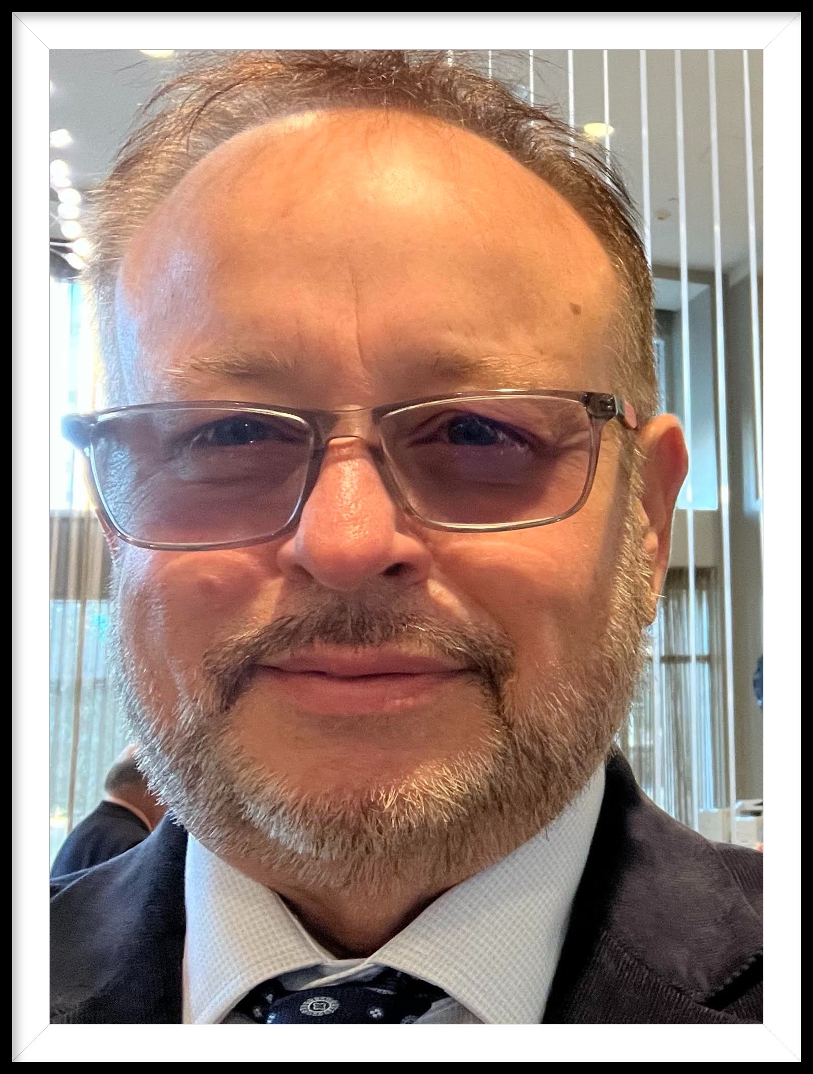 Man with glasses, beard, and blazer smiling, in a well-lit indoor setting.