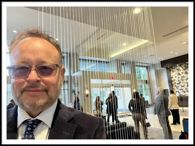 Man in a suit smiles, taking a selfie in a hotel lobby with vertical art and people in the background.