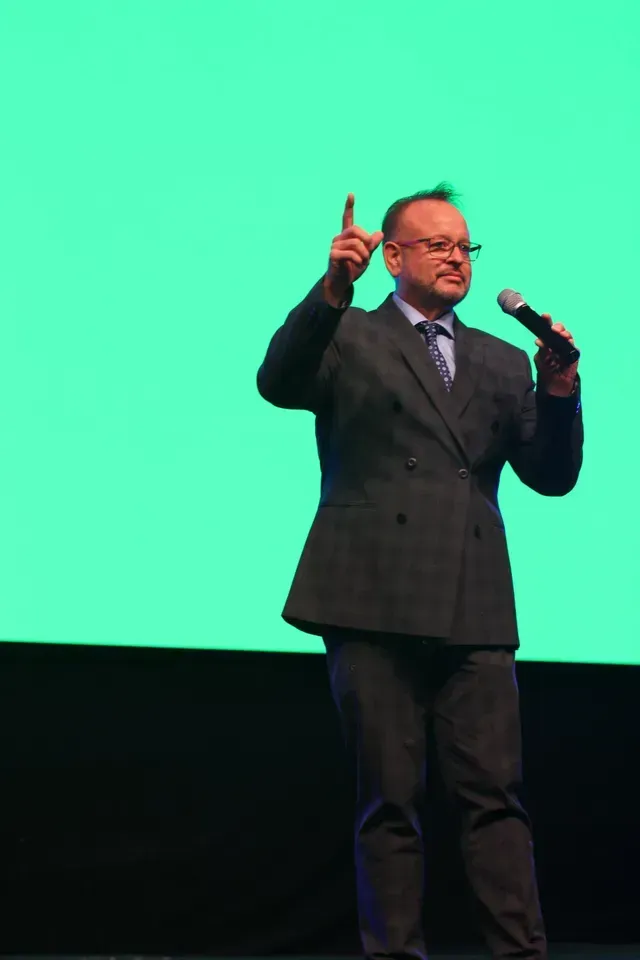 Man in suit speaking into a microphone, pointing upward with a bright green background.