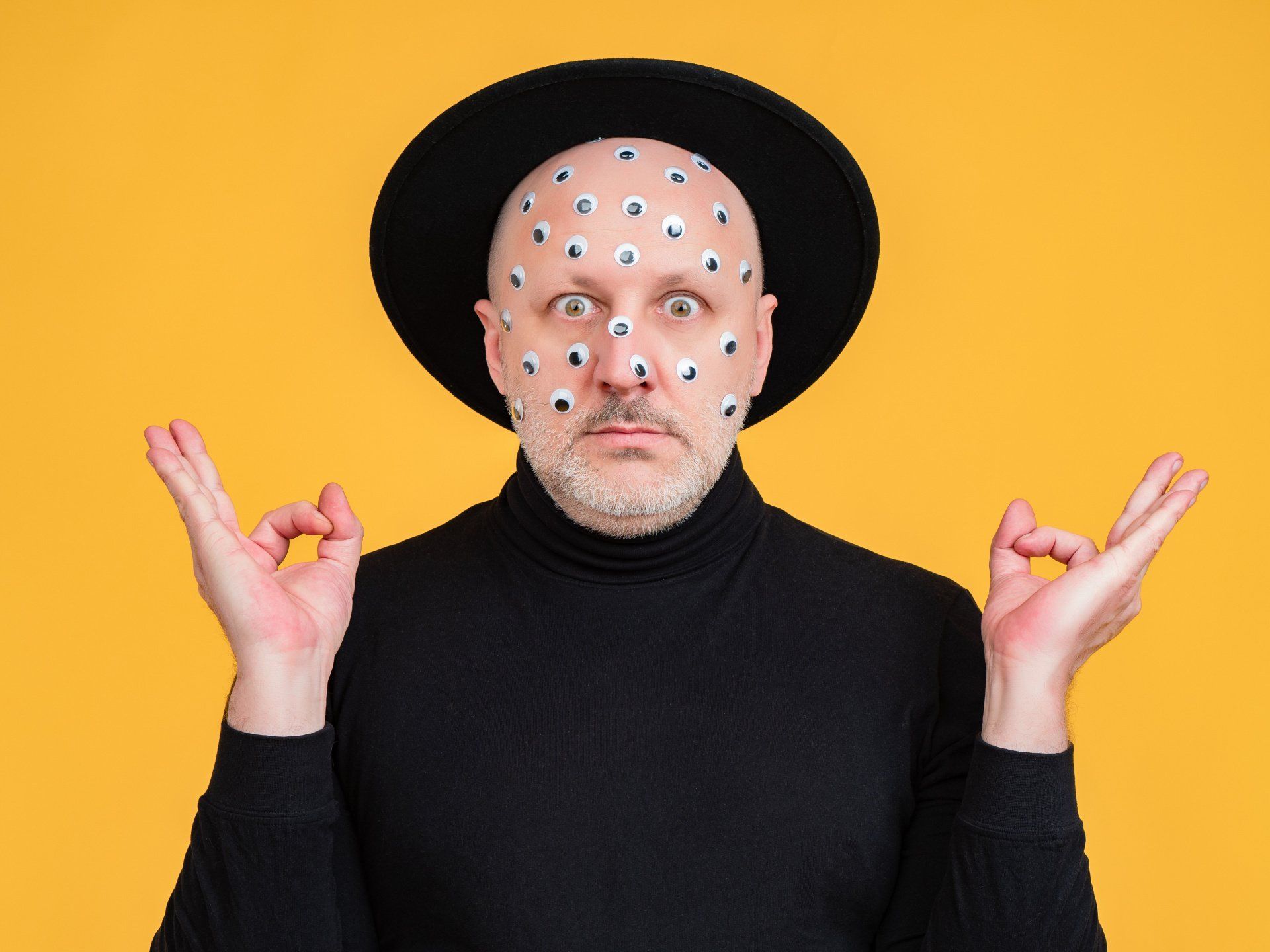 Man with eye stickers, black hat, and turtleneck; making an “okay” gesture against a yellow background.
