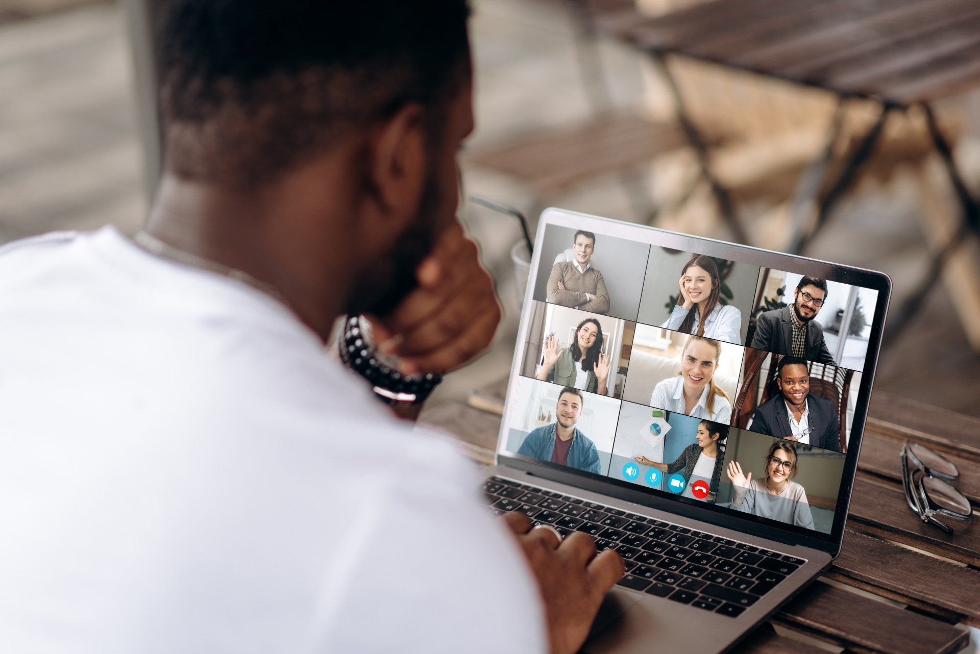 Person in white shirt attends a video call on a laptop outdoors. Several faces of meeting participants visible on screen.