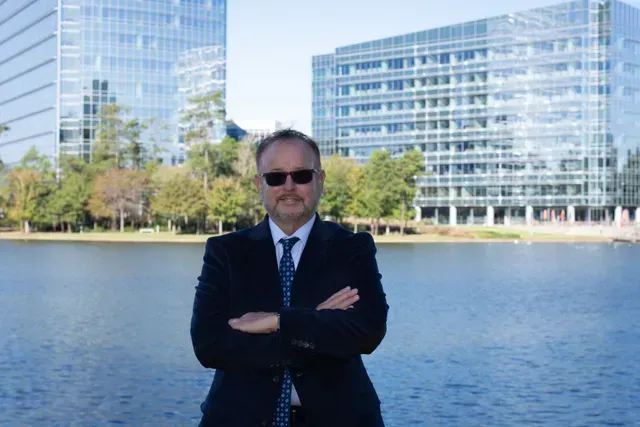 Man in suit with arms crossed, standing in front of a lake and buildings.