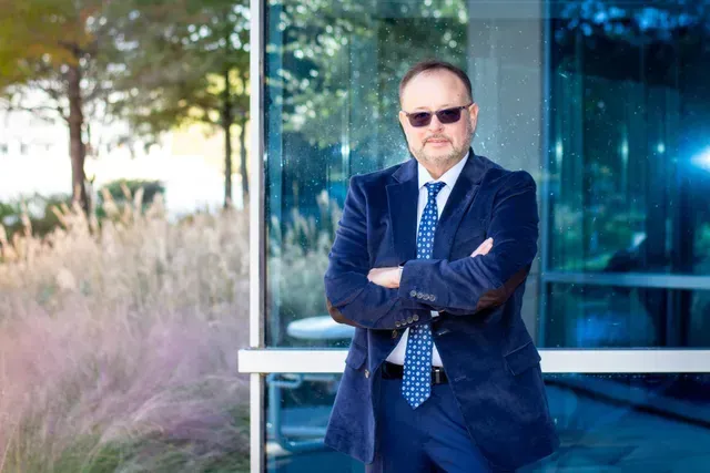 Man in sunglasses, blue suit, arms crossed, stands outside near glass building and plants.