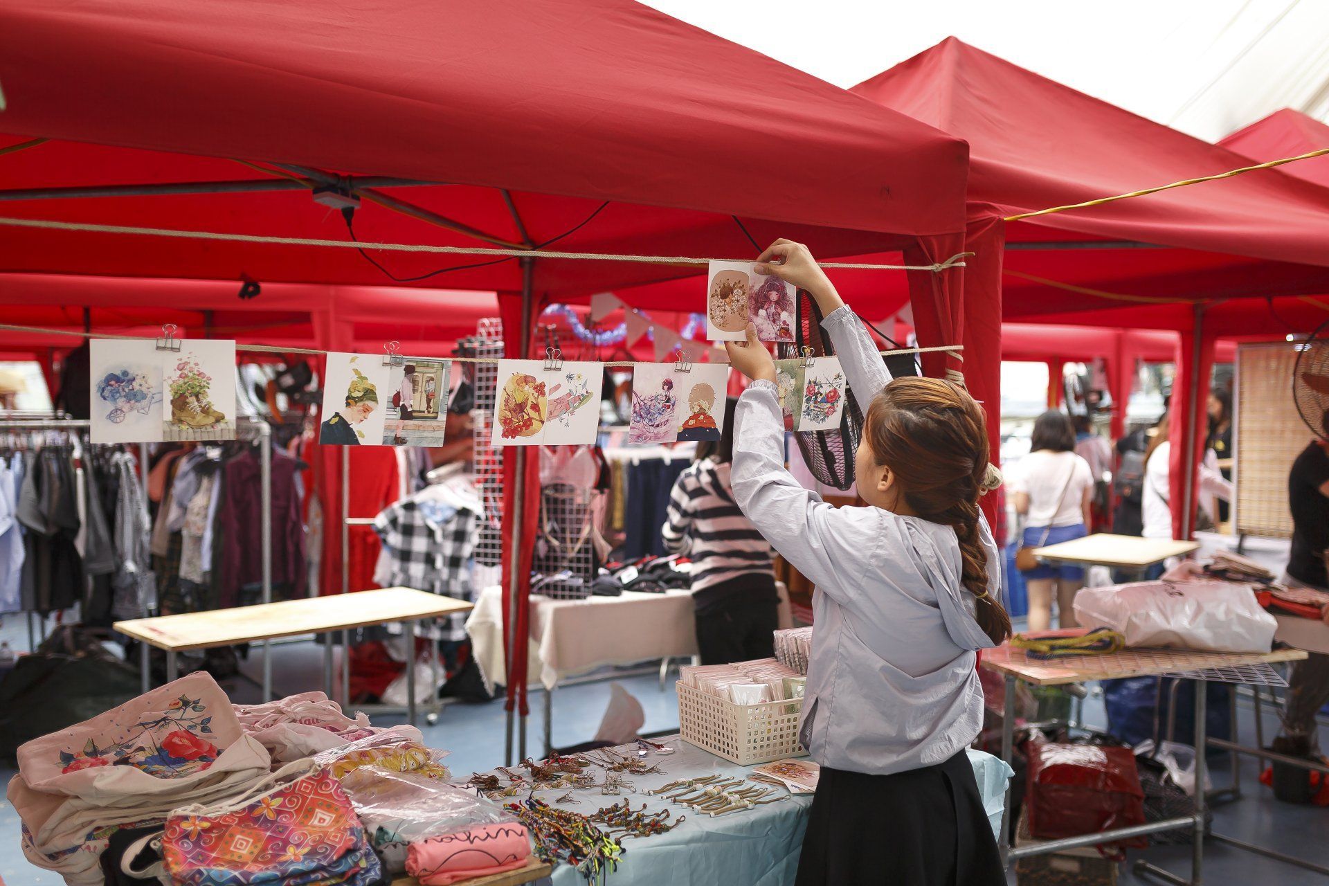 Woman arranging art prints at a red-tent market stall. Merchandise includes clothing, bags, and jewelry.
