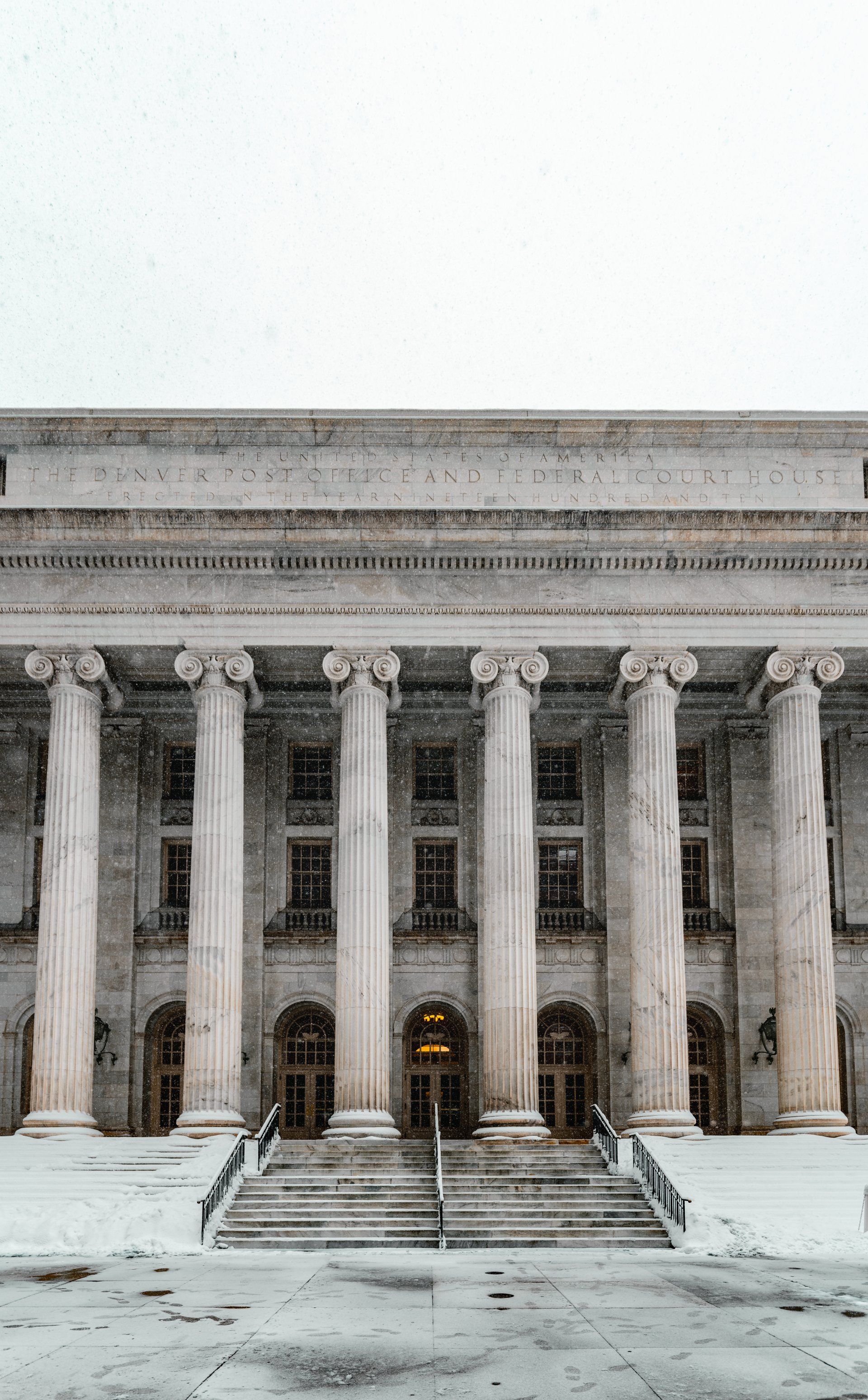A classical building with tall columns, snow falling on it, and a snowy ground.