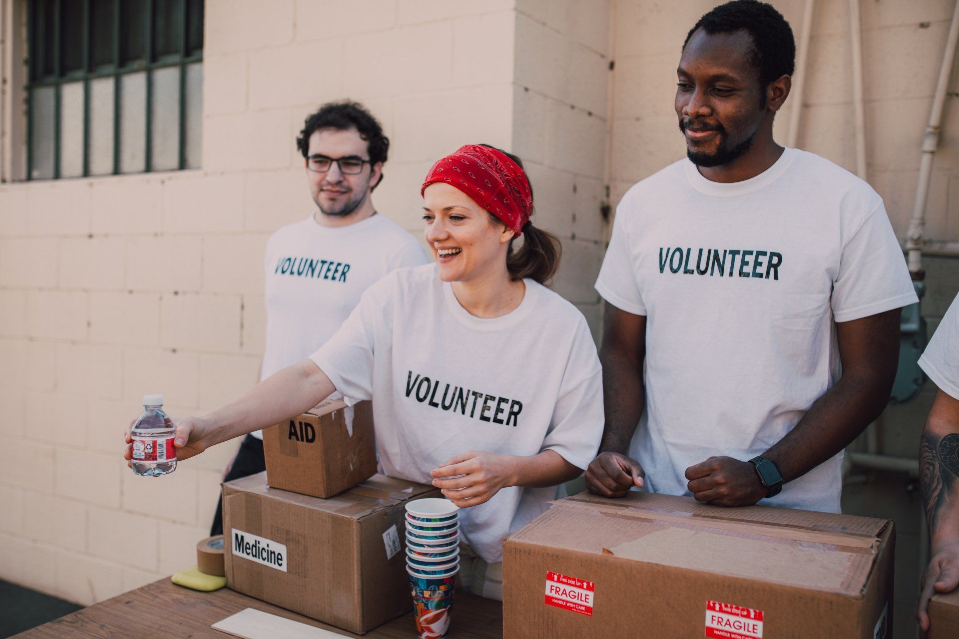 Volunteers at a table handing out water bottles and supplies, wearing white shirts with "VOLUNTEER" printed on them.