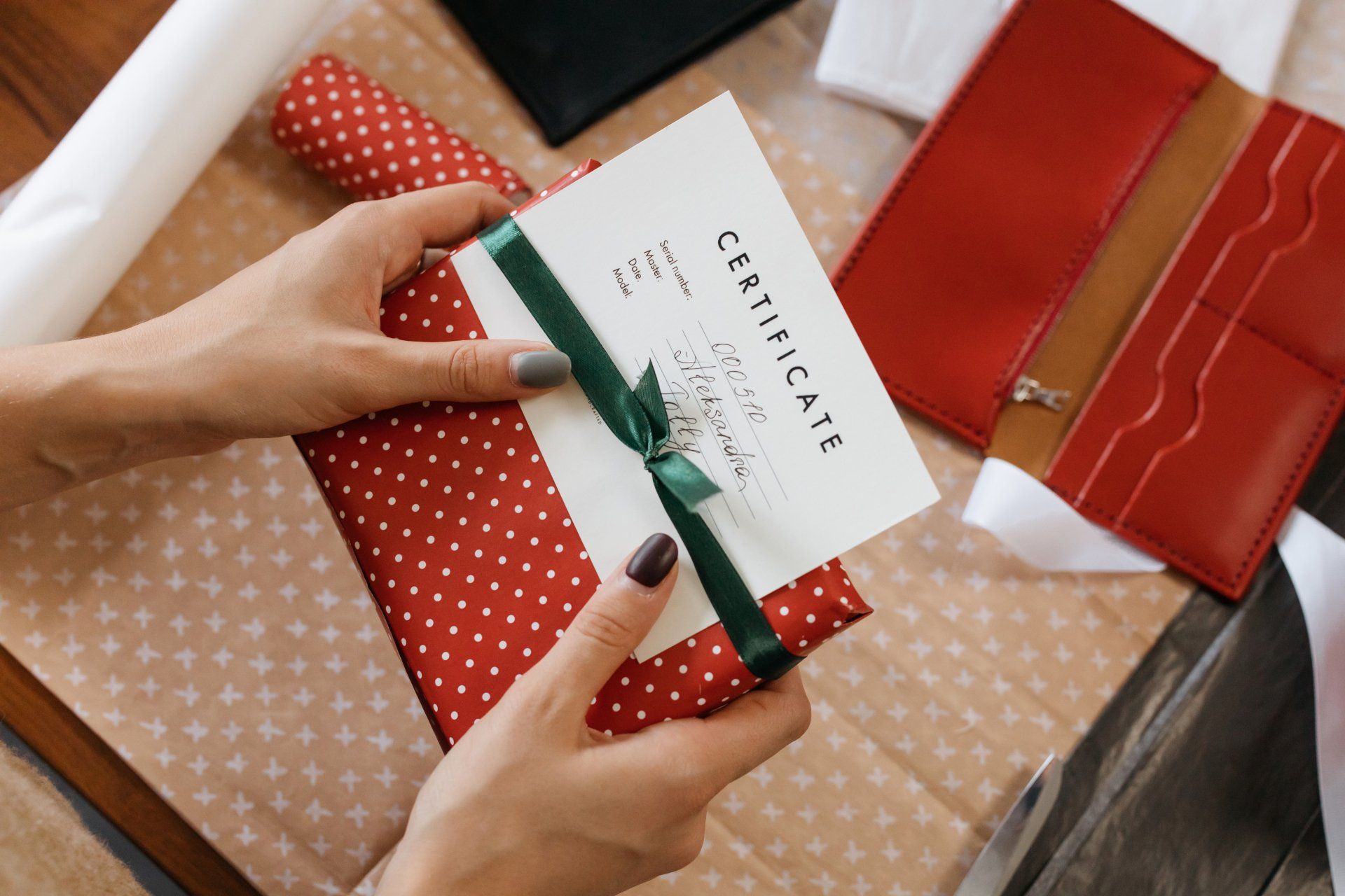 Hands holding a wrapped gift with a green ribbon and a certificate. Red, white, and brown items in the background.