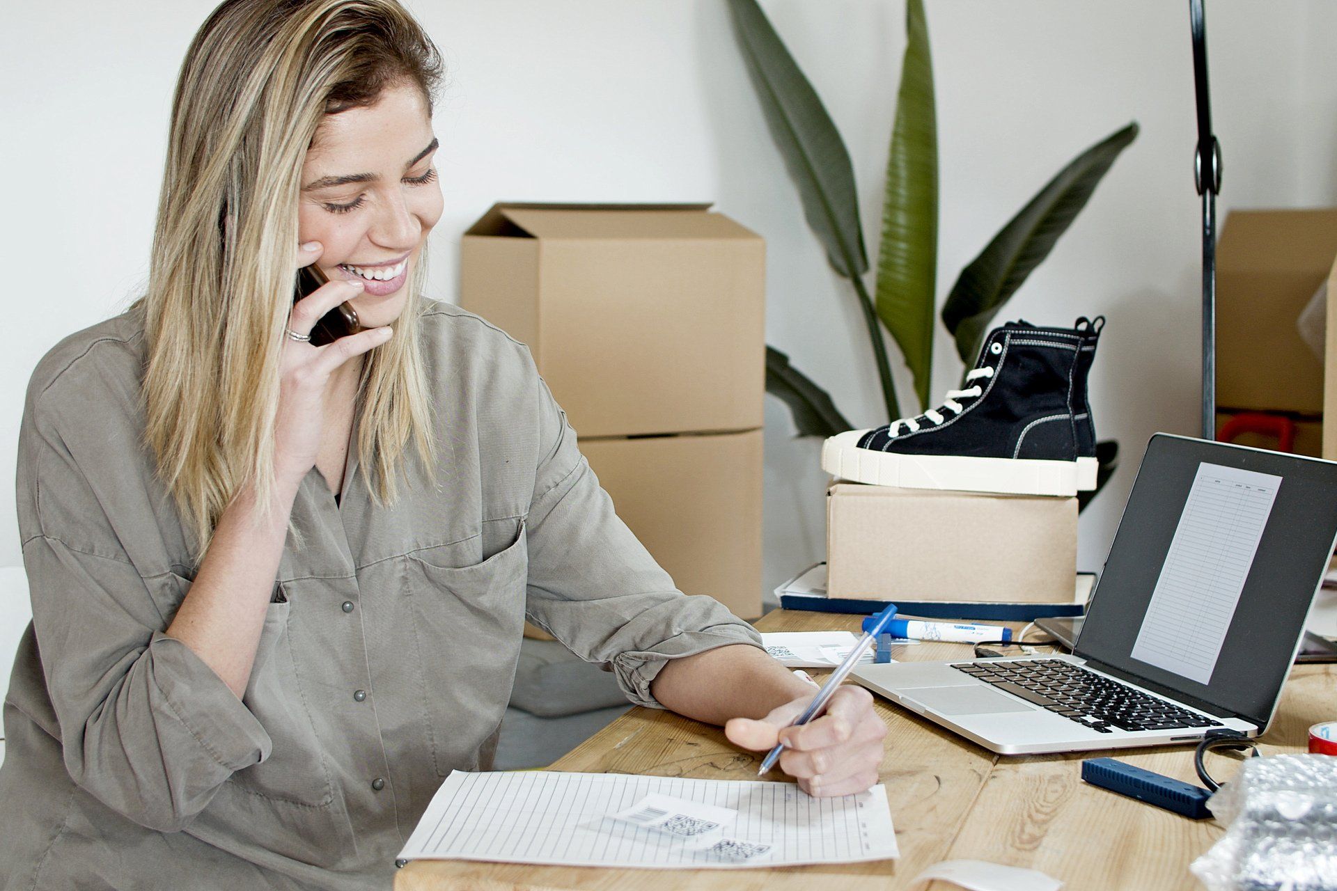 Woman on phone, smiling, writing at desk with laptop, shoe, boxes.