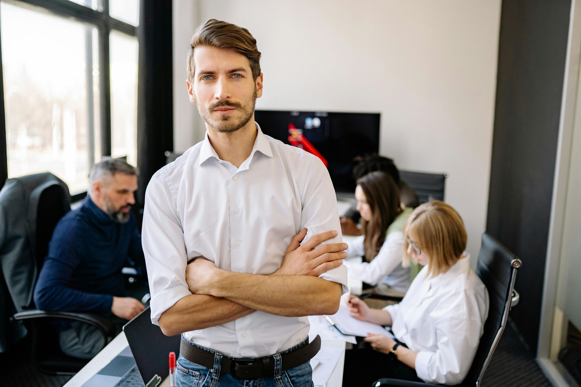 Man with crossed arms in office, other people in background at a table.