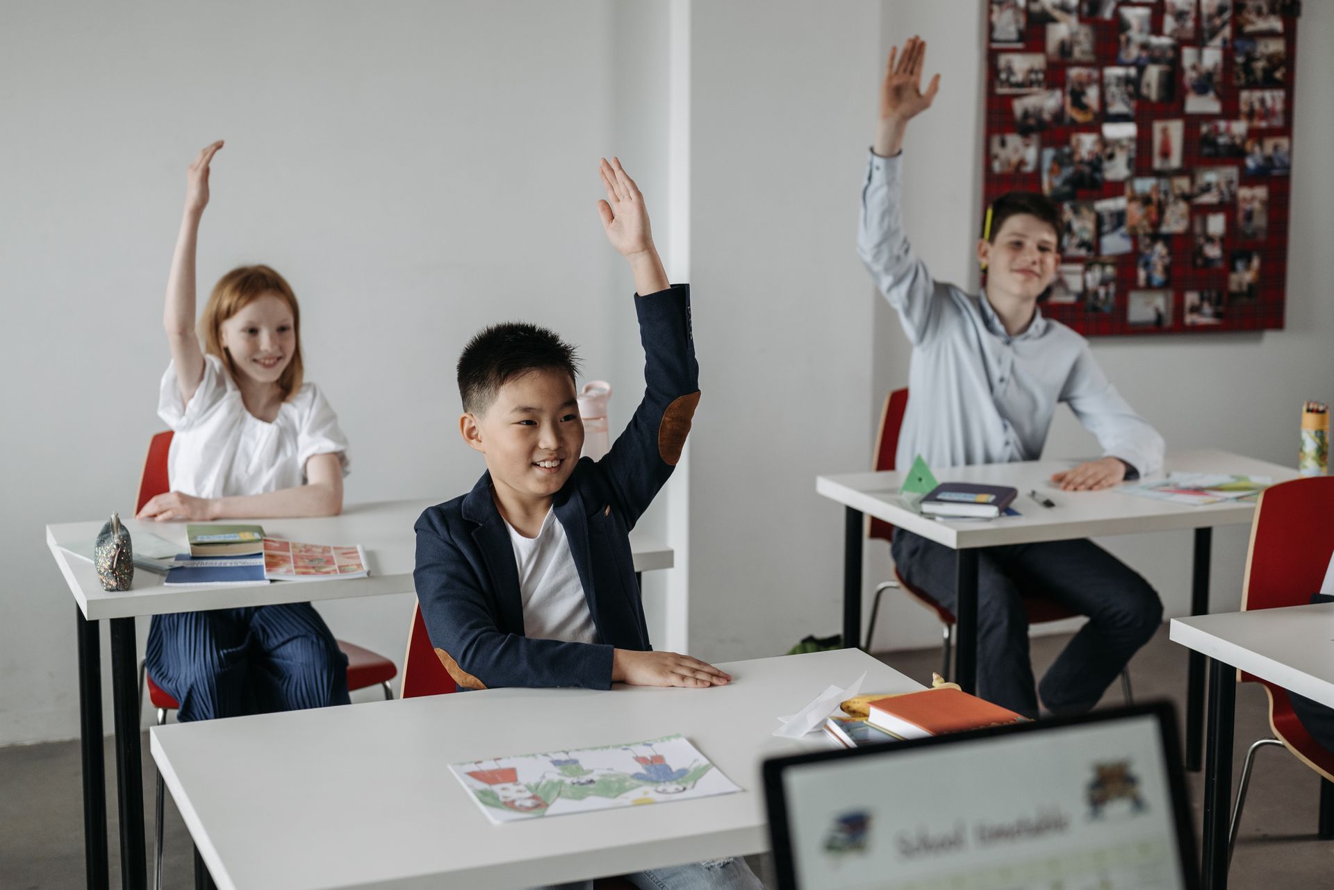 Three students raising their hands in a classroom, smiling at their desks.