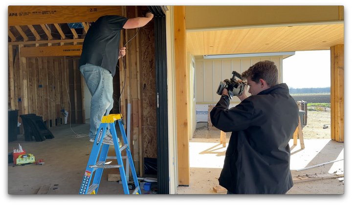 A man on a ladder is working on a door while another man takes a picture with a camera.