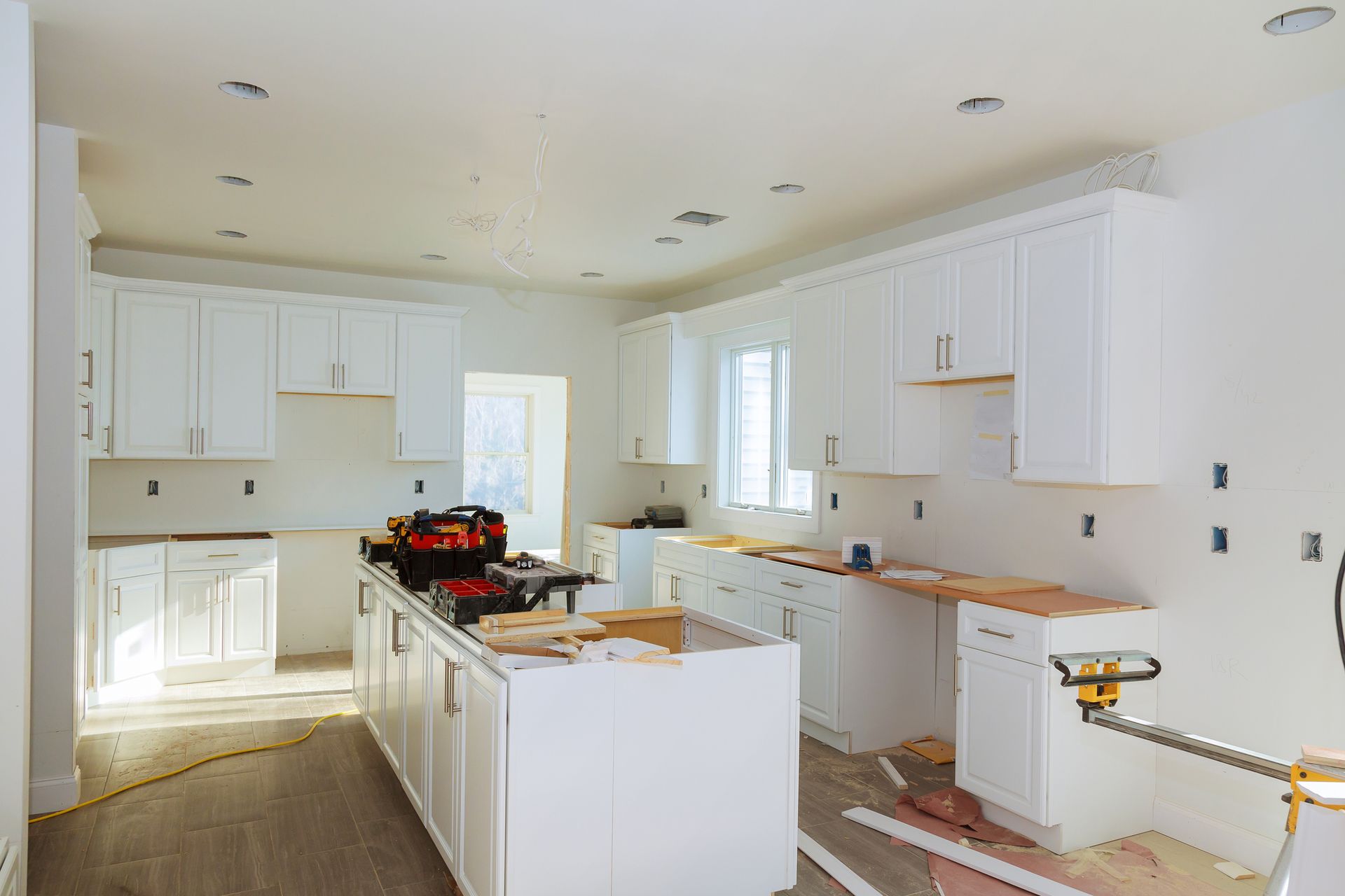 White kitchen remodel in progress; cabinets installed, tools on island, light walls and wood floor.
