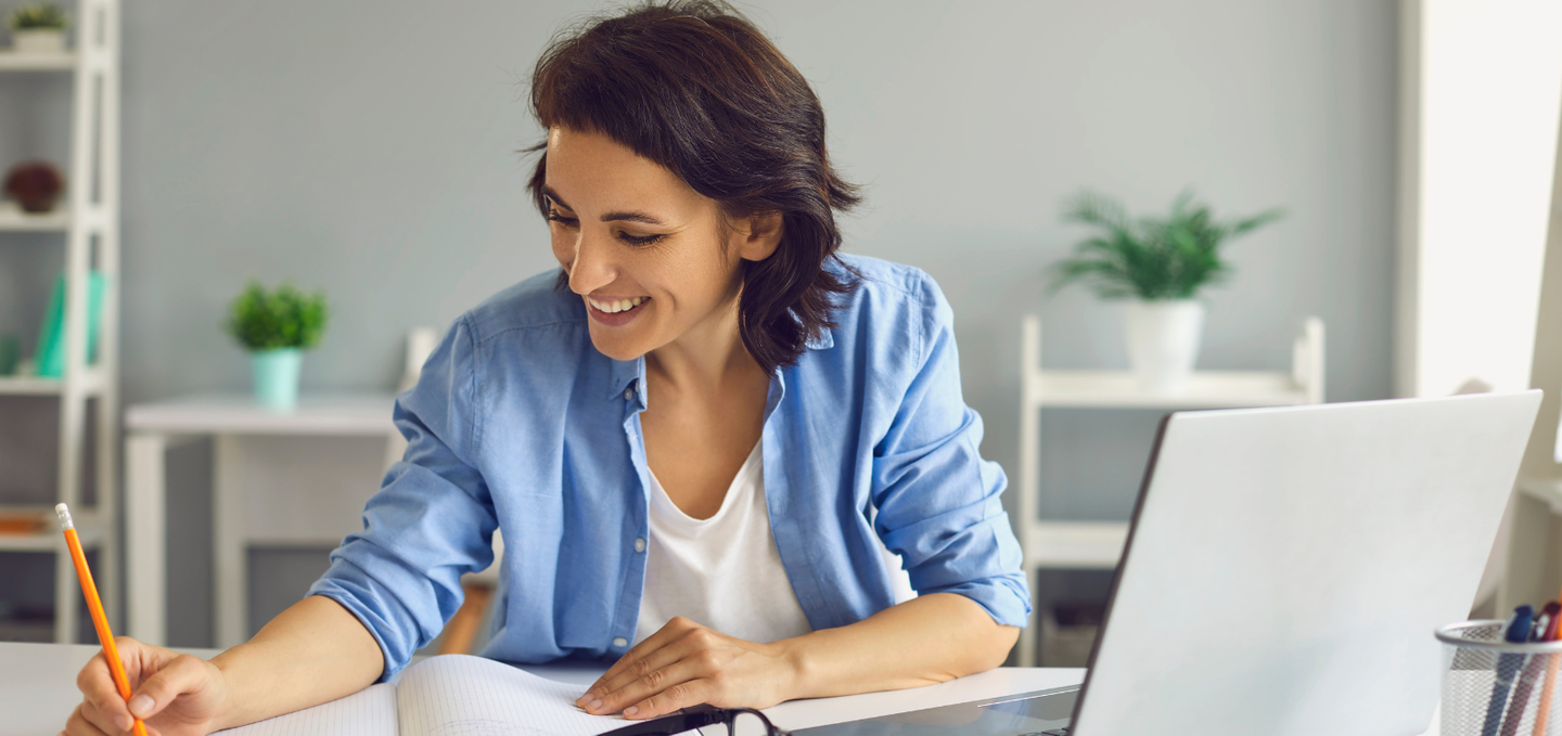 A woman is sitting at a desk with a laptop and writing in a notebook.