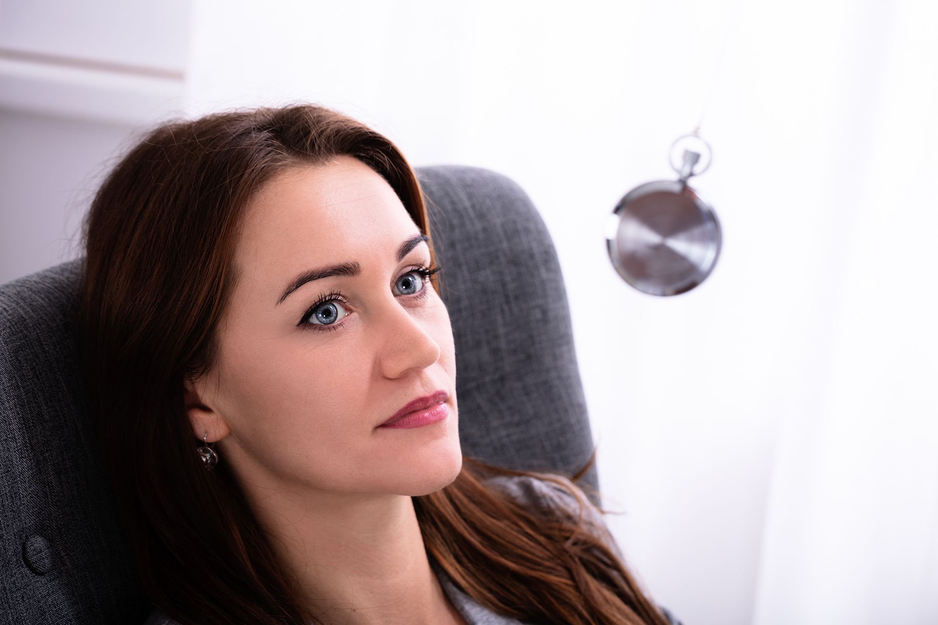 Woman gazing at a swinging pocket watch, seated in a chair, possibly in a clinical setting.
