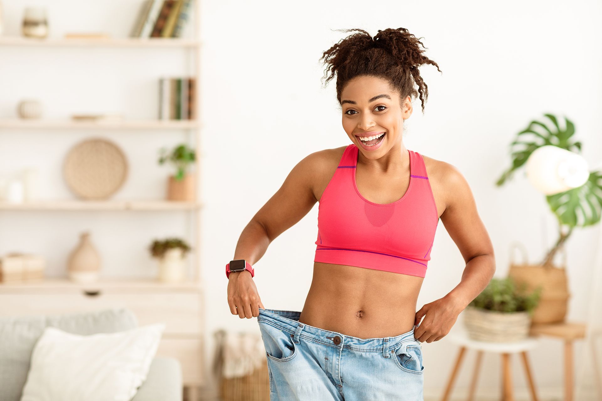 Woman smiling, wearing oversized jeans, showing weight loss, standing in a bright room.