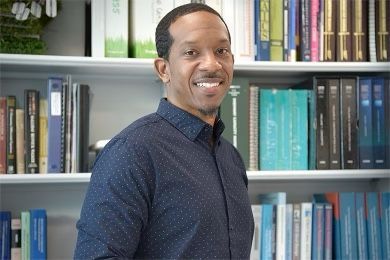 Man with dark skin, smiling in front of a bookshelf filled with books and binders. He wears a dark blue button-down shirt.