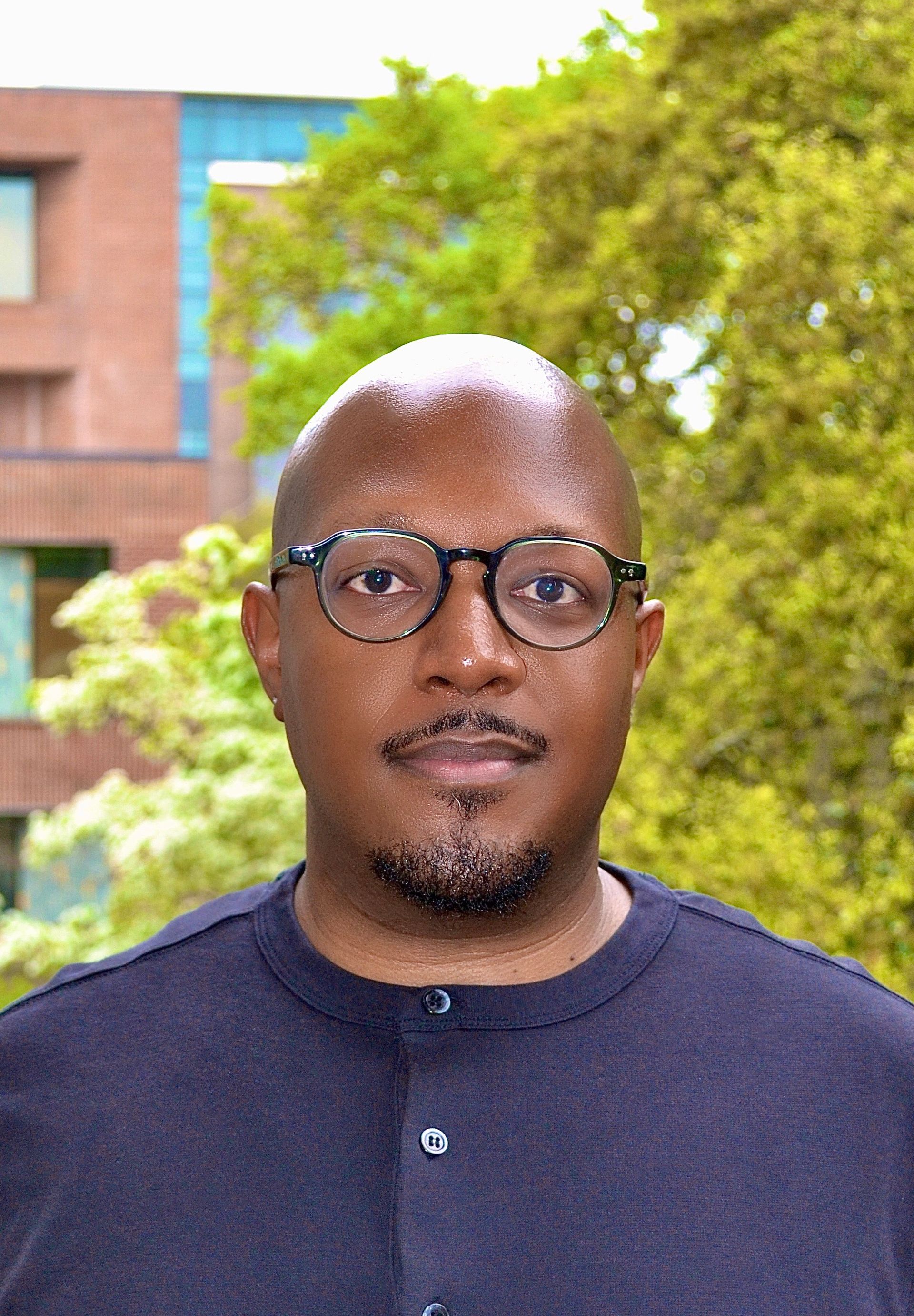 Bald Black man with glasses in front of a building and green foliage, wearing a dark button-up shirt, looking at the camera.
