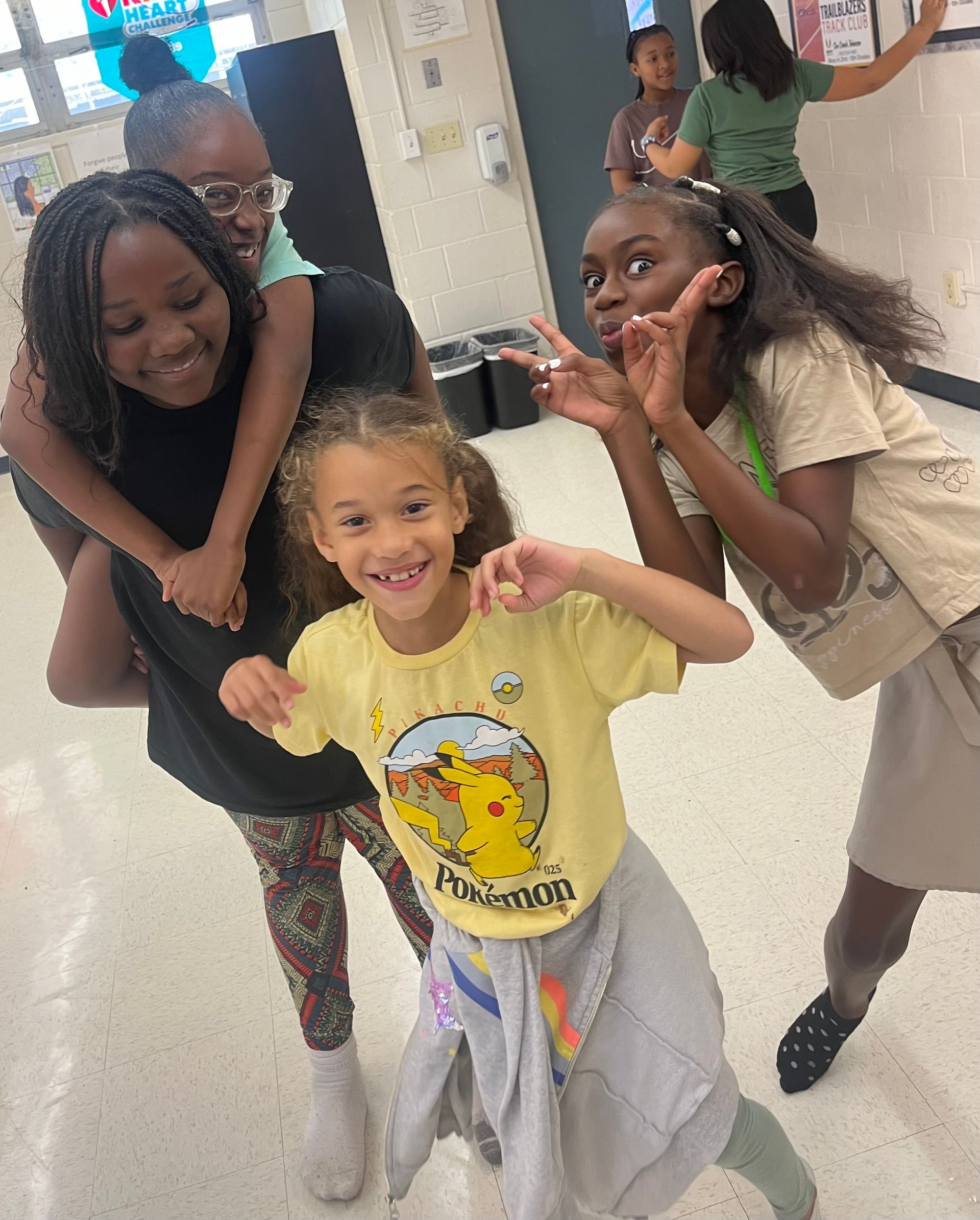 A group of diverse kids smiling and posing in a hallway. One girl is piggybacking another, while another makes a peace sign.