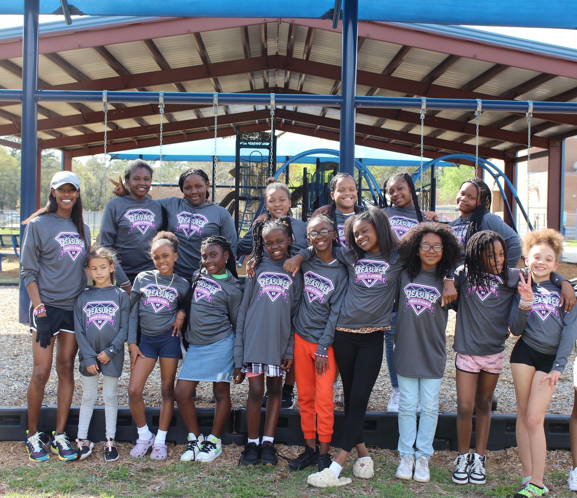 A youth cheerleading team in gray shirts with pink accents poses in front of a playground. They smile and embrace each other.
