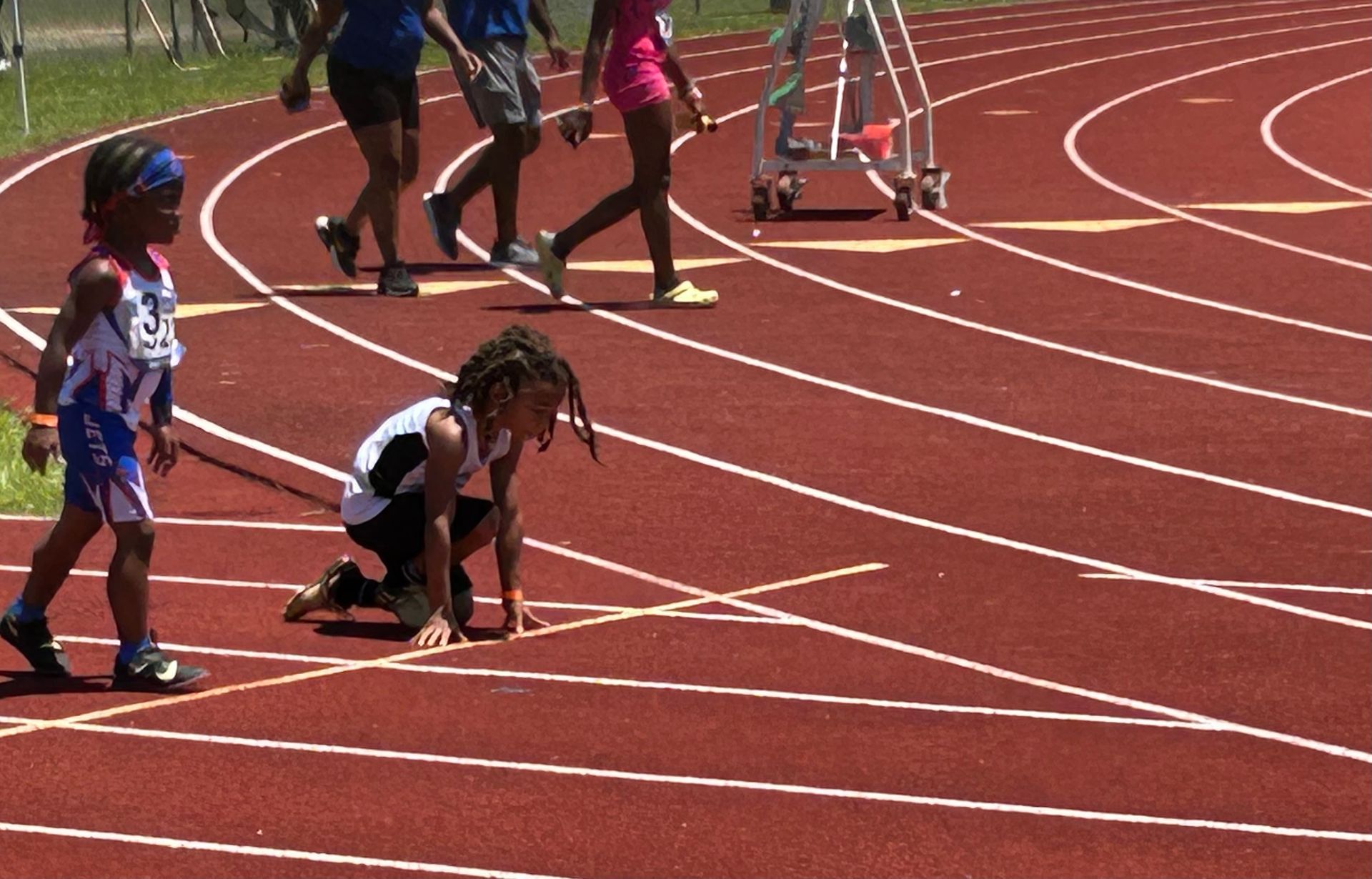 Young athlete crouches at the starting line of a track race, preparing to run. Other runners walk the track in the background.