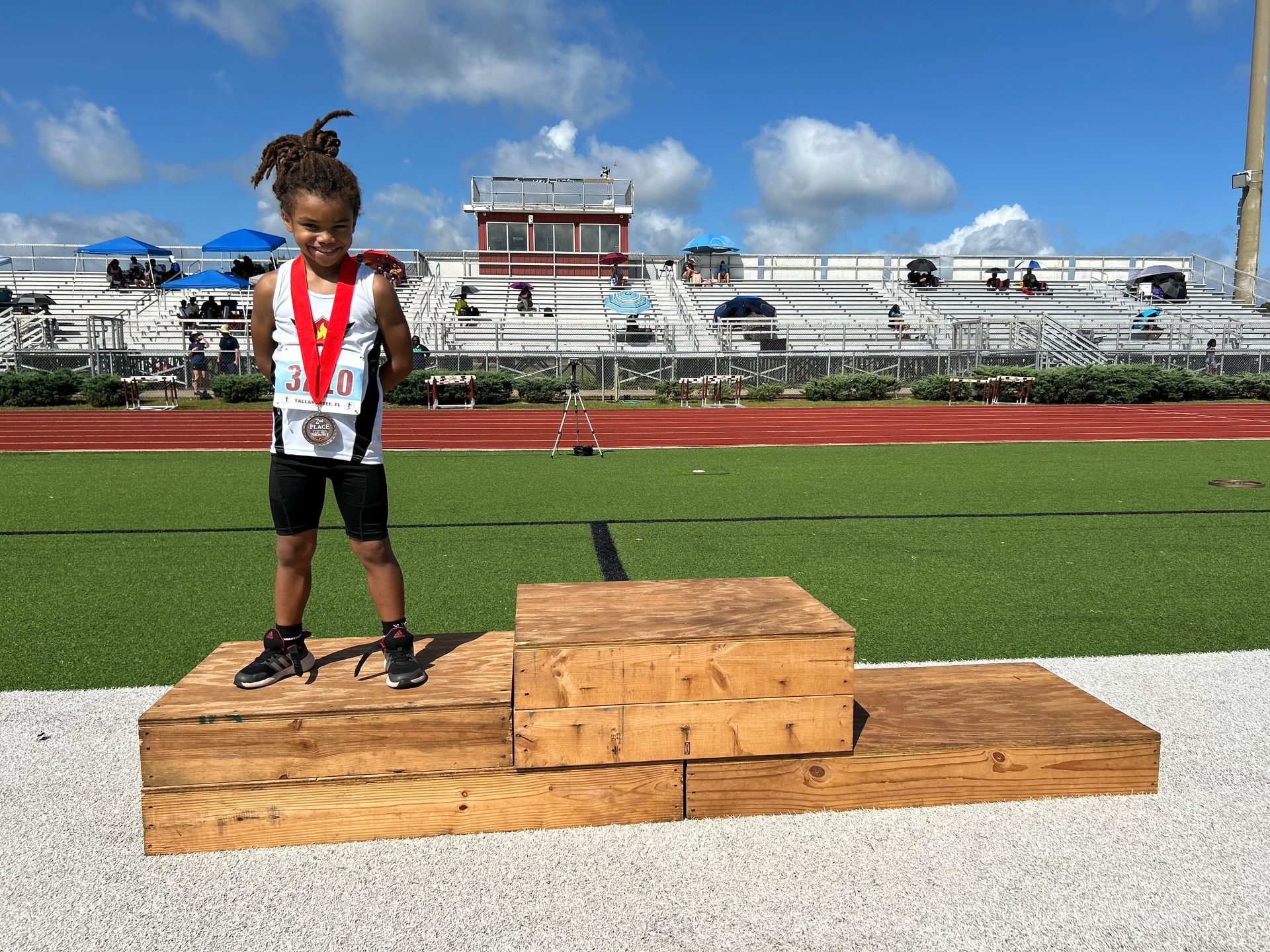 Young athlete with dreadlocks stands on a wooden podium, wearing a medal, on a track field.