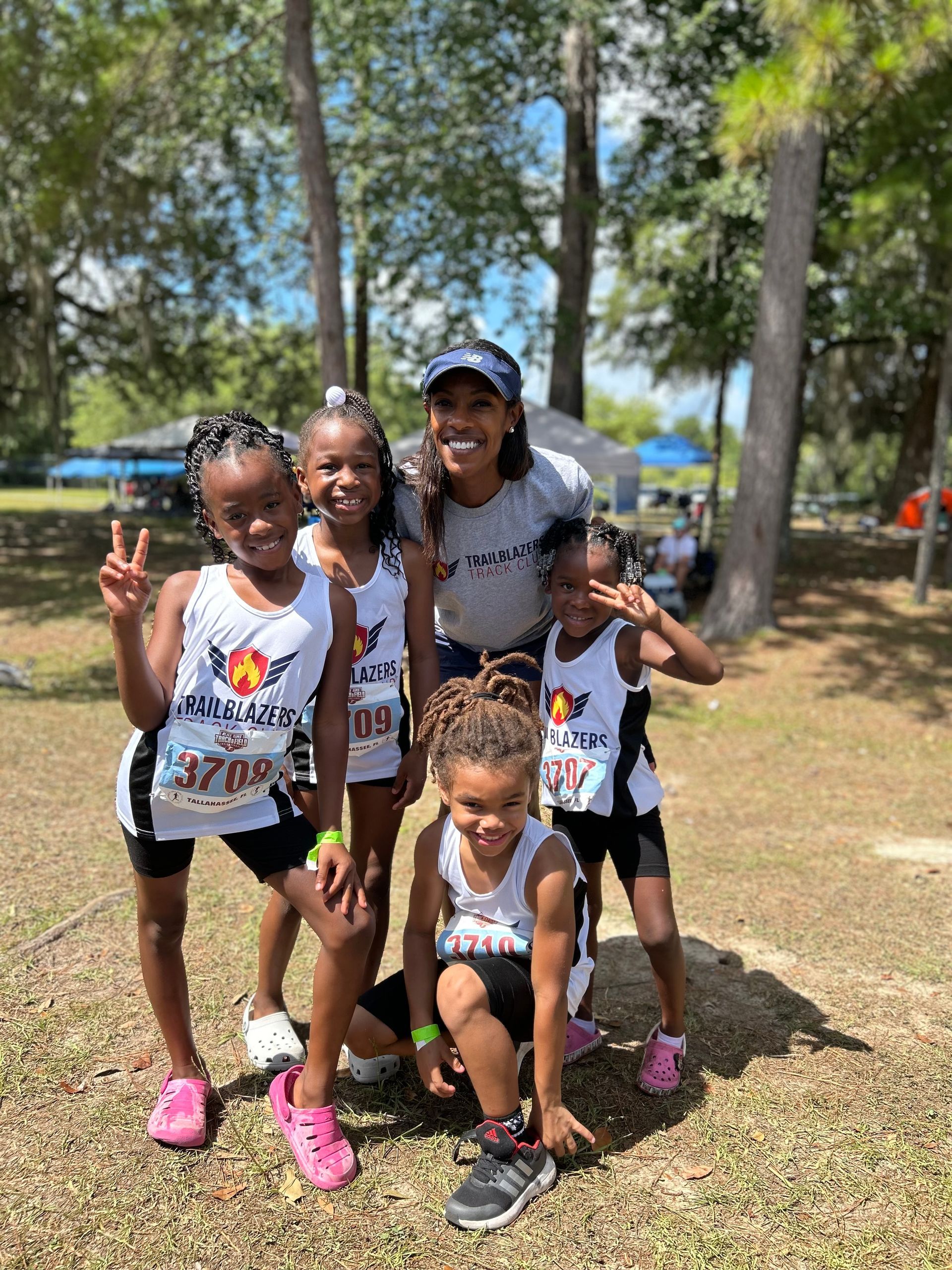 A group of young Black girls and an adult woman pose in athletic gear outdoors. Some are smiling and giving peace signs.