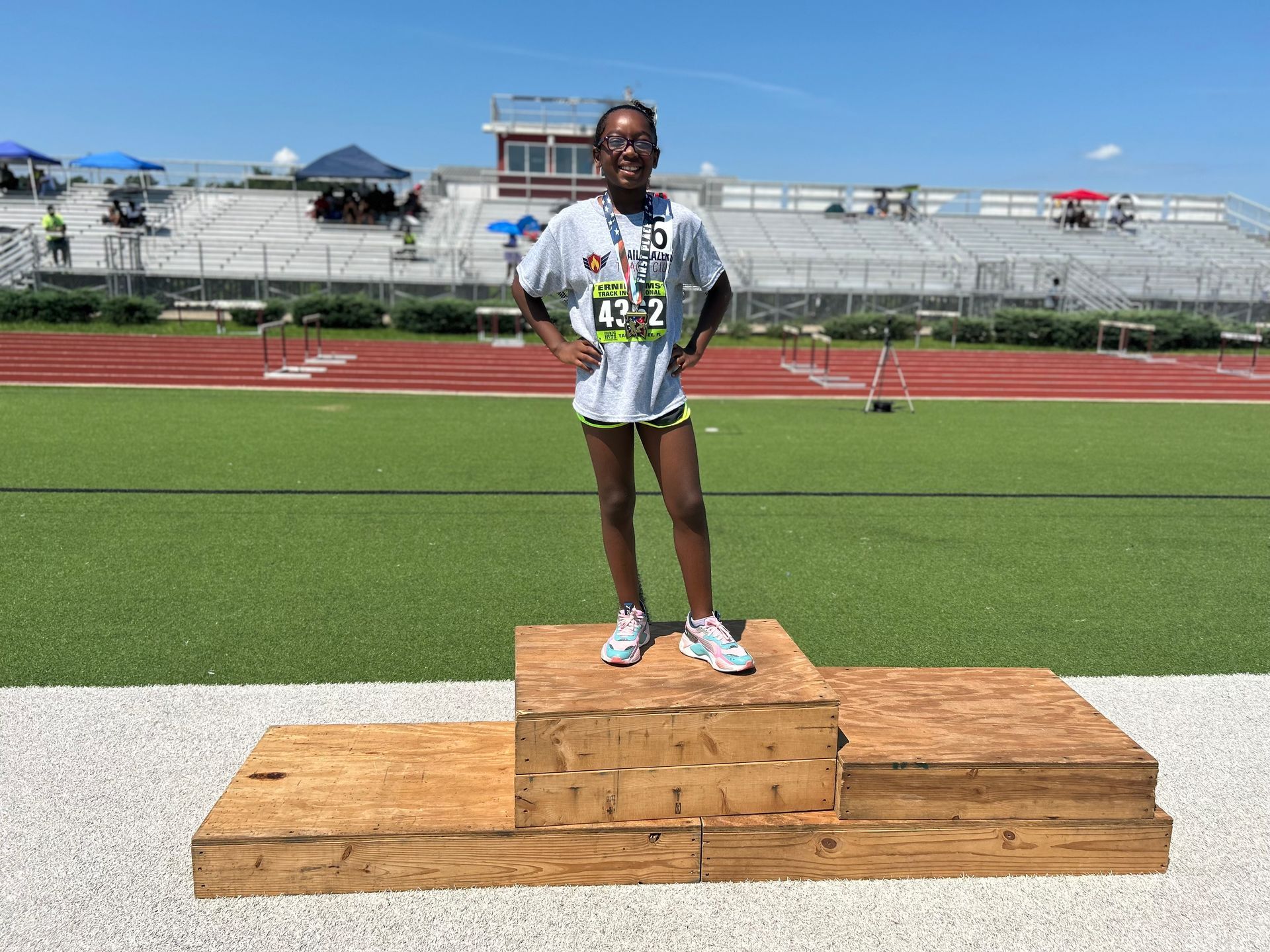 A young girl stands on a podium at a track and field event, wearing a medal. She is smiling, outdoors on a sunny day.