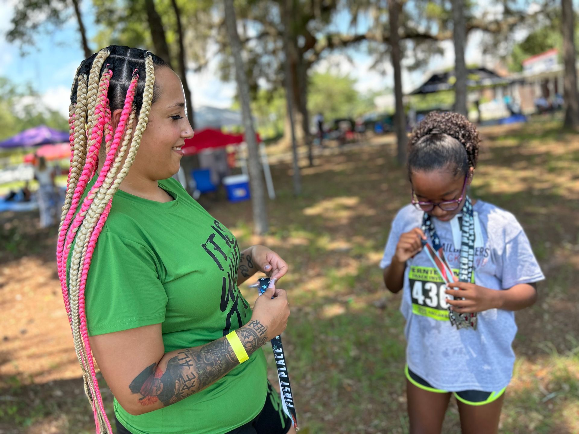 Woman with pink and blonde braids and a young girl with glasses, in a park, holding medals after a race.