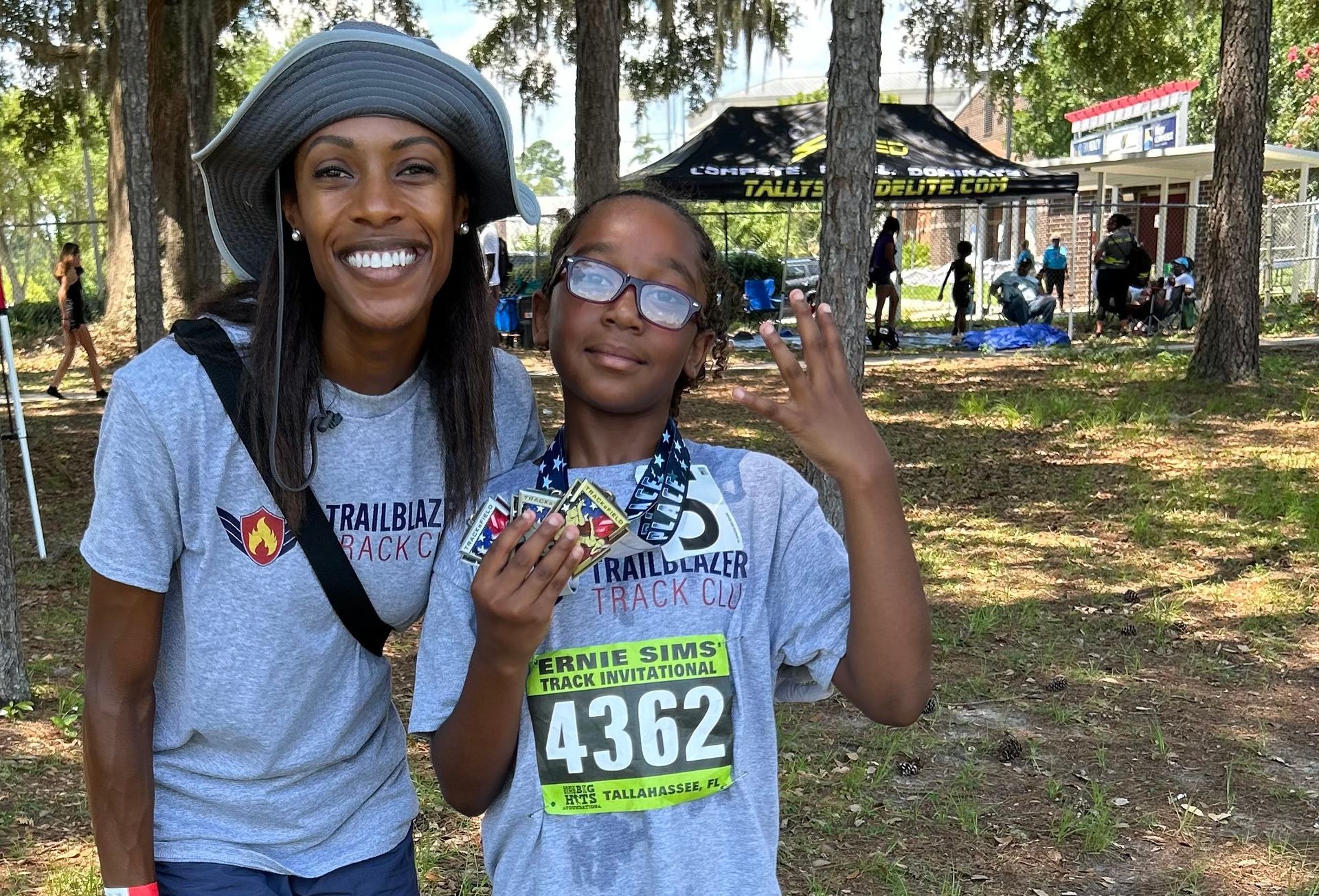 Woman and child smiling at a race. The child holds a medal, wearing a race bib. Setting is outdoors, with trees and a tent in the background.