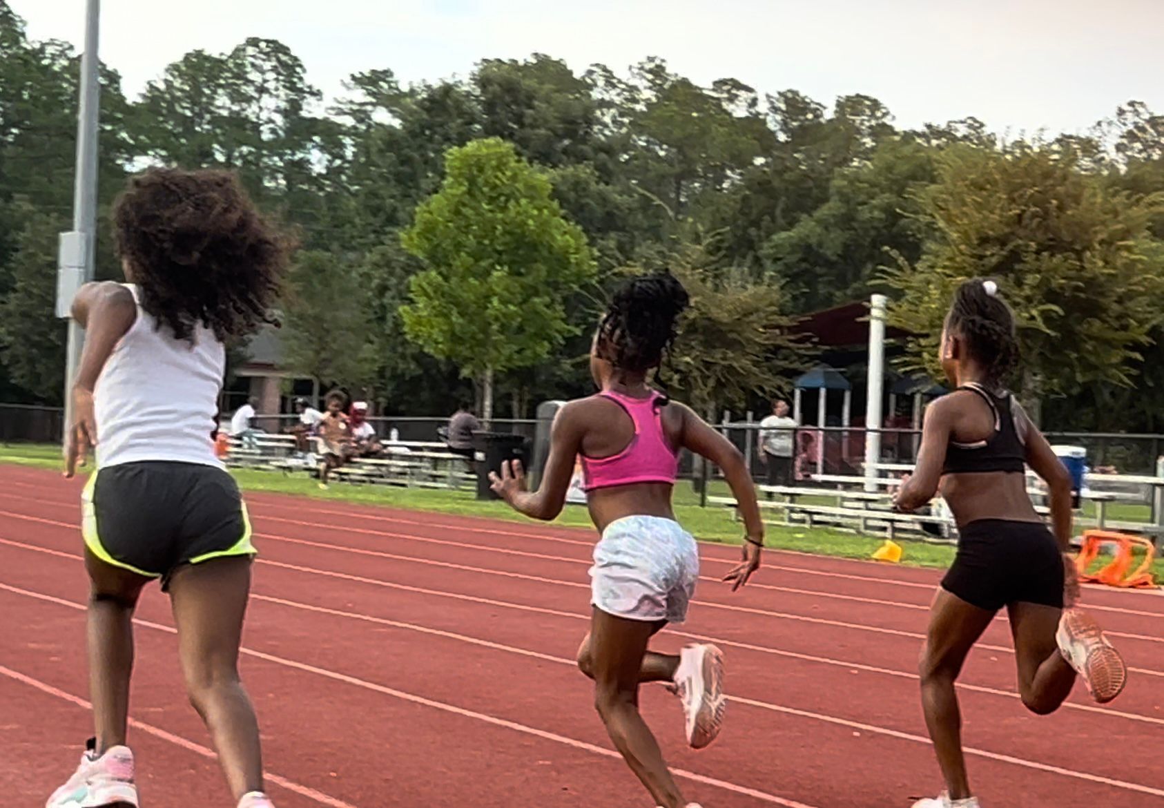 Three young girls running on a track. They wear athletic clothes: a white tank top and yellow shorts, pink sports bra and white shorts, and black sports bra and shorts.