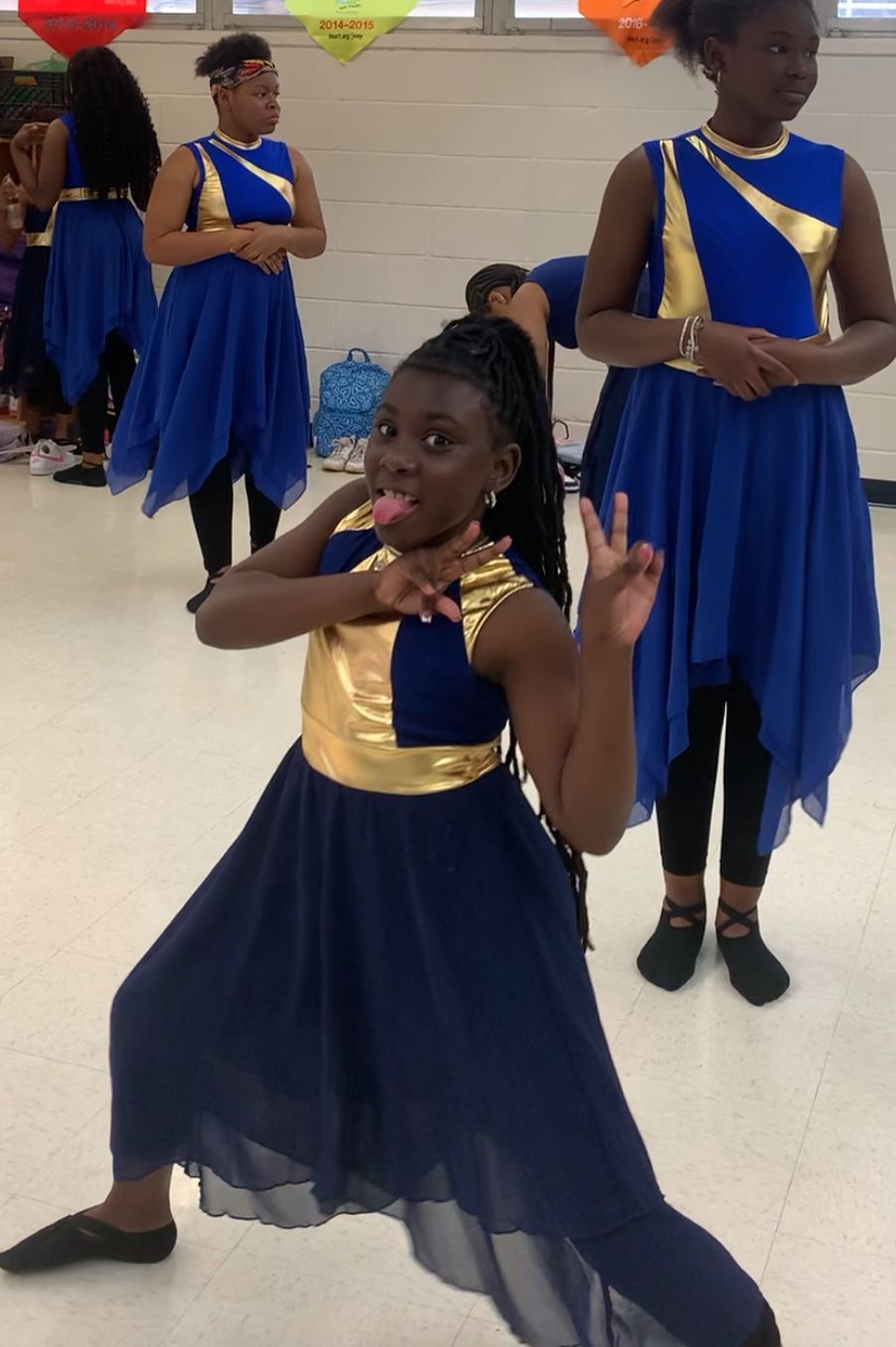 A young Black girl in a blue and gold dance costume poses playfully, flanked by other dancers in a school setting.