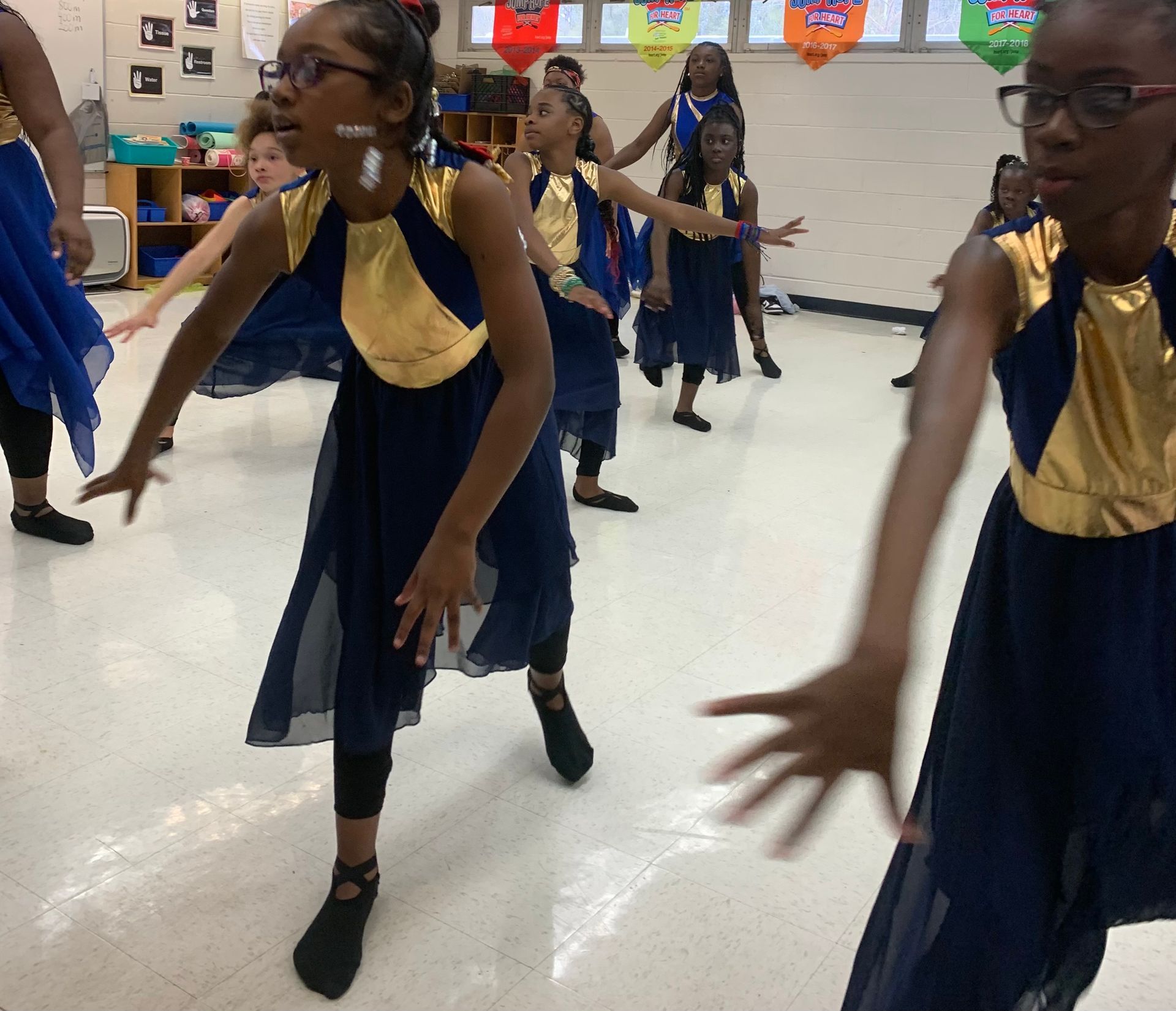 Group of young dancers in blue and gold dresses performing in a classroom. They have their arms outstretched.