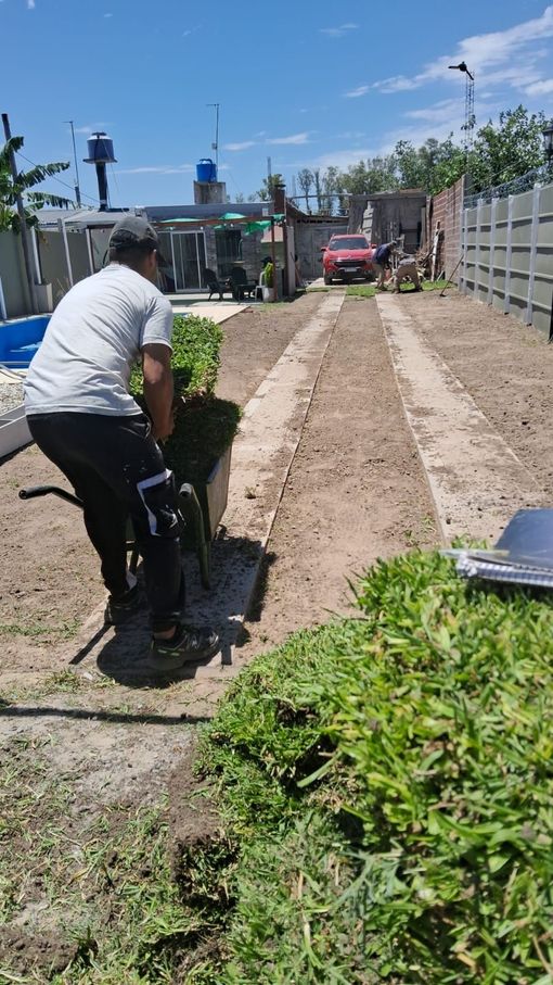 Hombre usando una carretilla para mover tierra en un patio, camión rojo en la distancia.