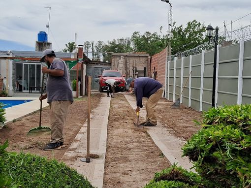 Dos personas rastrillando y preparando un cantero entre caminos de hormigón en un patio trasero.