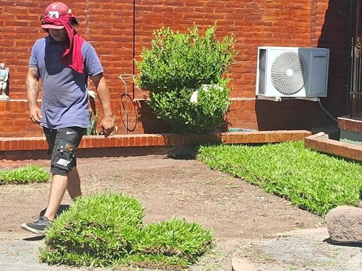 Hombre trabajando en jardinería, colocando césped. Al fondo, una pared de ladrillo rojo, un aire acondicionado y un arbusto verde.