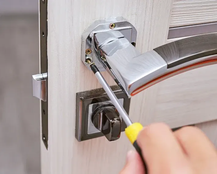 A person is using a screwdriver to install a shiny silver door handle on a light wooden door.