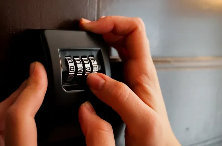 Person's hands turning dials on a black combination lock attached to a brown door, showing numbers 6226.