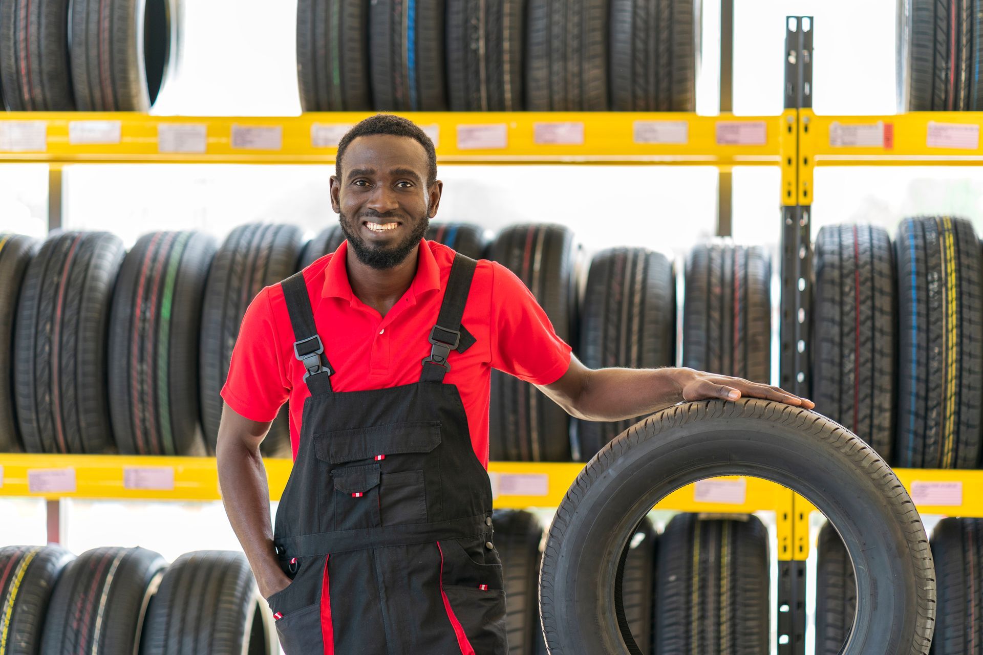 Mechanic in red shirt and overalls smiles, leaning on tire in a tire shop, tires on shelves behind. Mechanic in red shirt and overalls smiles, leaning on tire in a tire shop, tires on shelves behind.