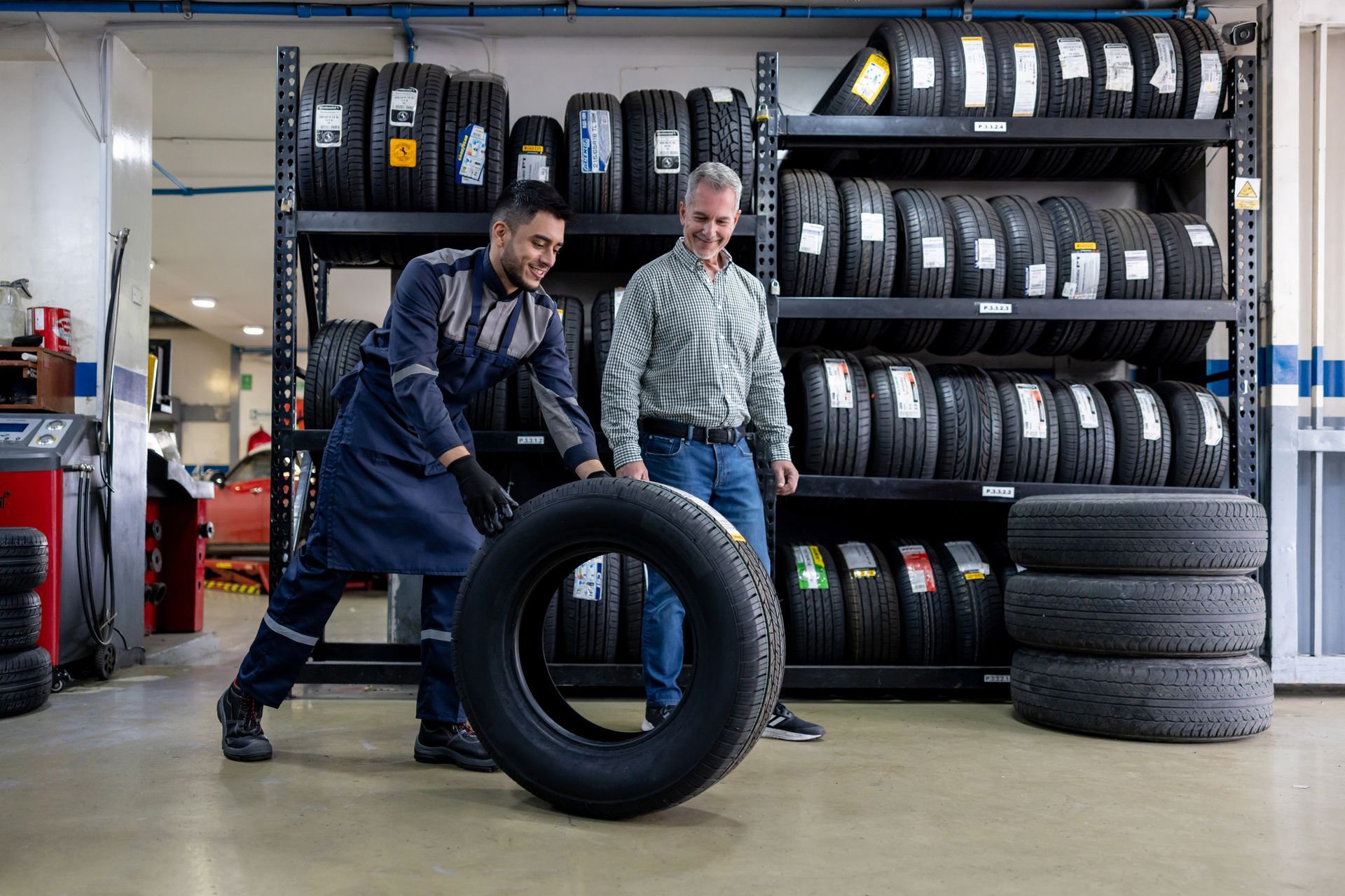 A mechanic, in a blue uniform, is moving a tire and talking to his client at a used tire shop.