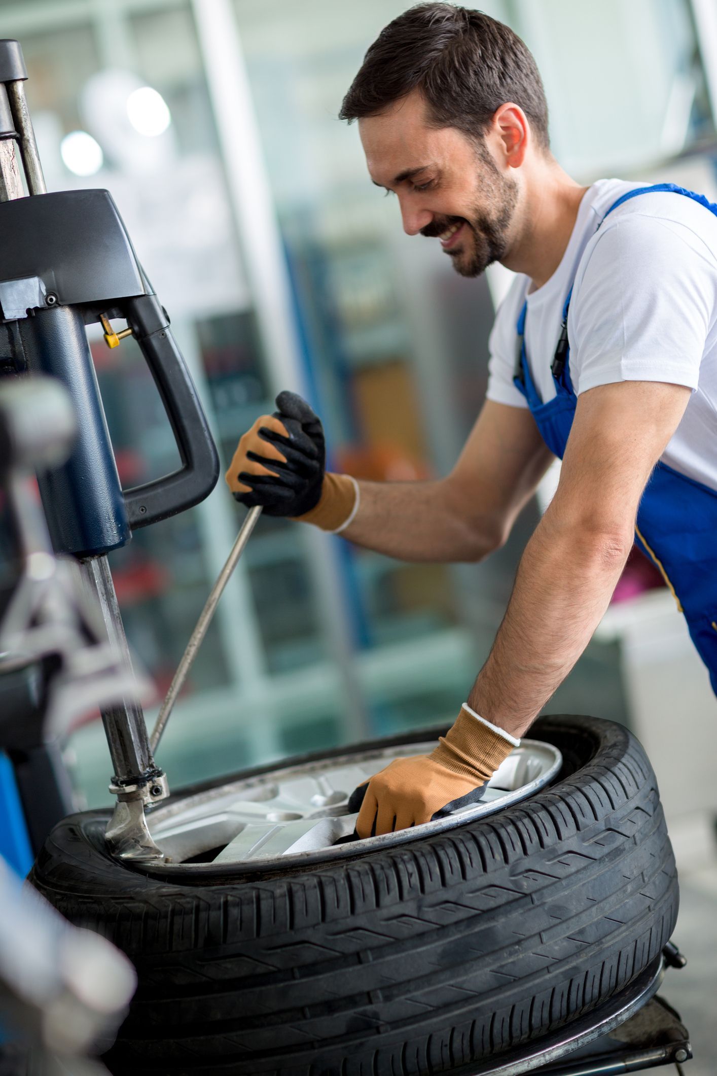 Mechanic using tool to mount tire on rim in a shop. He is wearing gloves and smiling.
