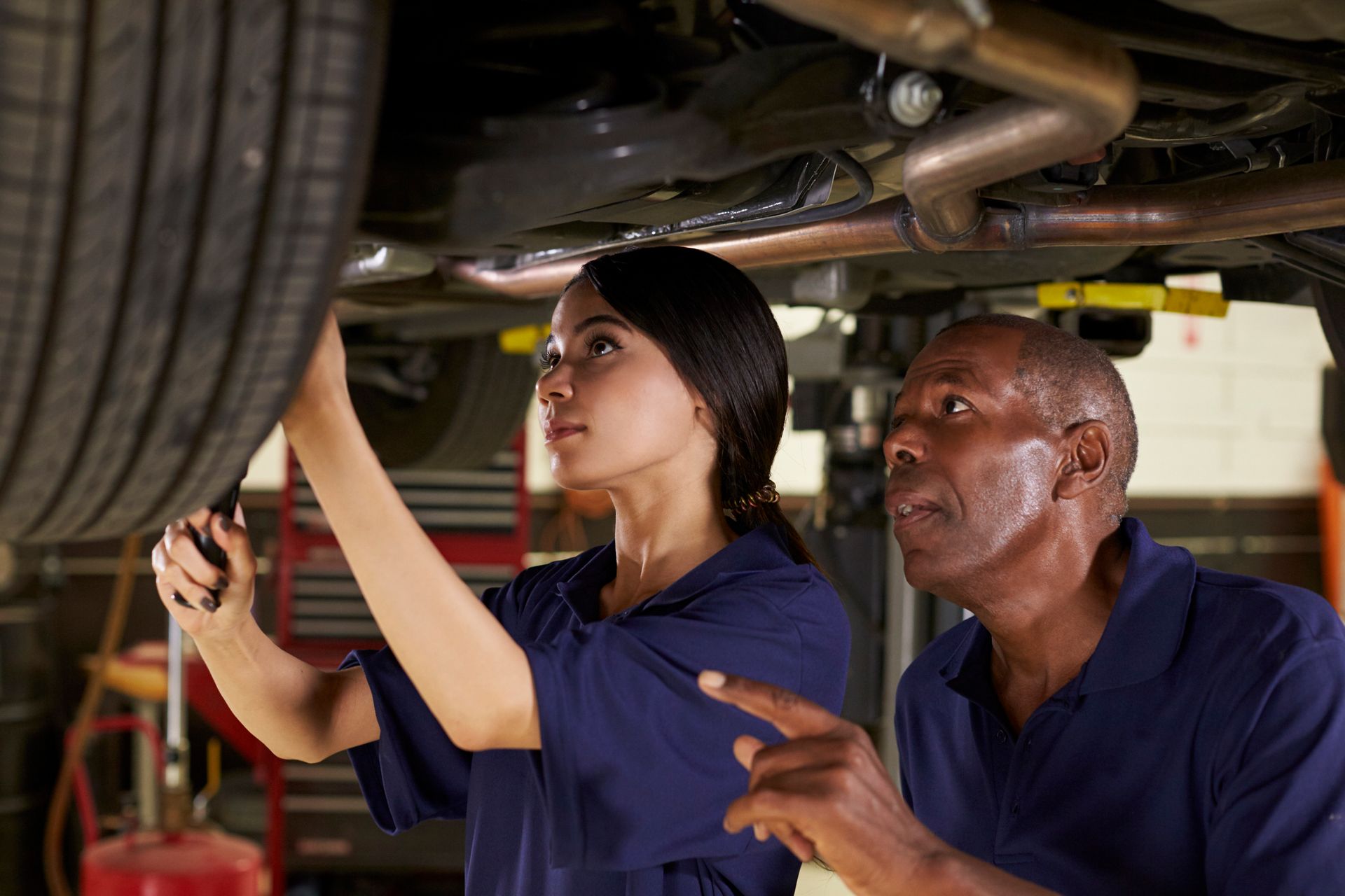 Mechanic points at vehicle components as a colleague works on the undercarriage in a garage.