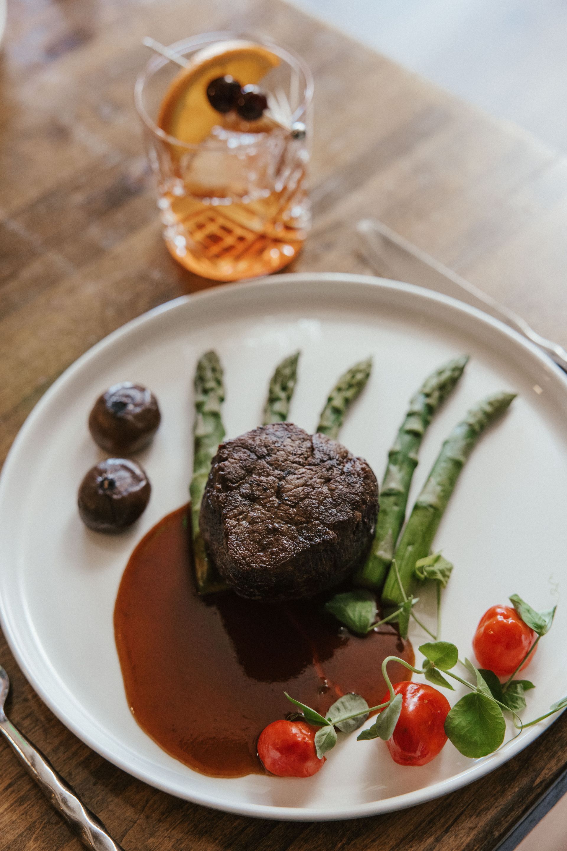 A white plate topped with a steak , asparagus , tomatoes and sauce on a wooden table.