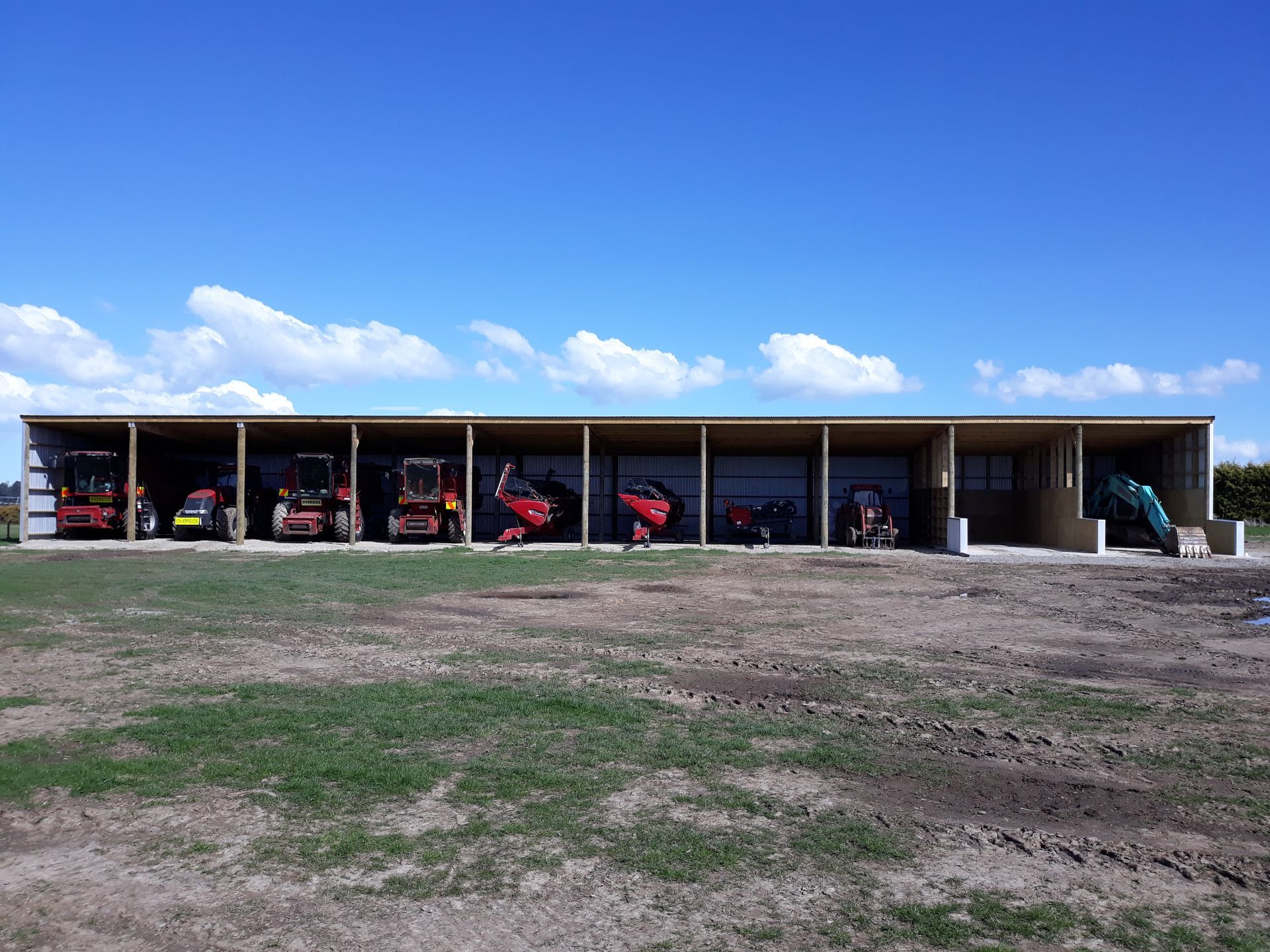 A row of tractors are parked under a shed in a field.
