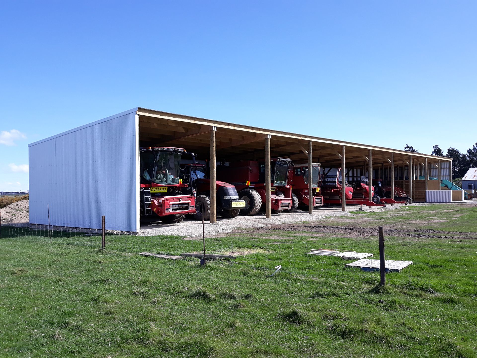 A row of tractors are parked under a shed in a grassy field.