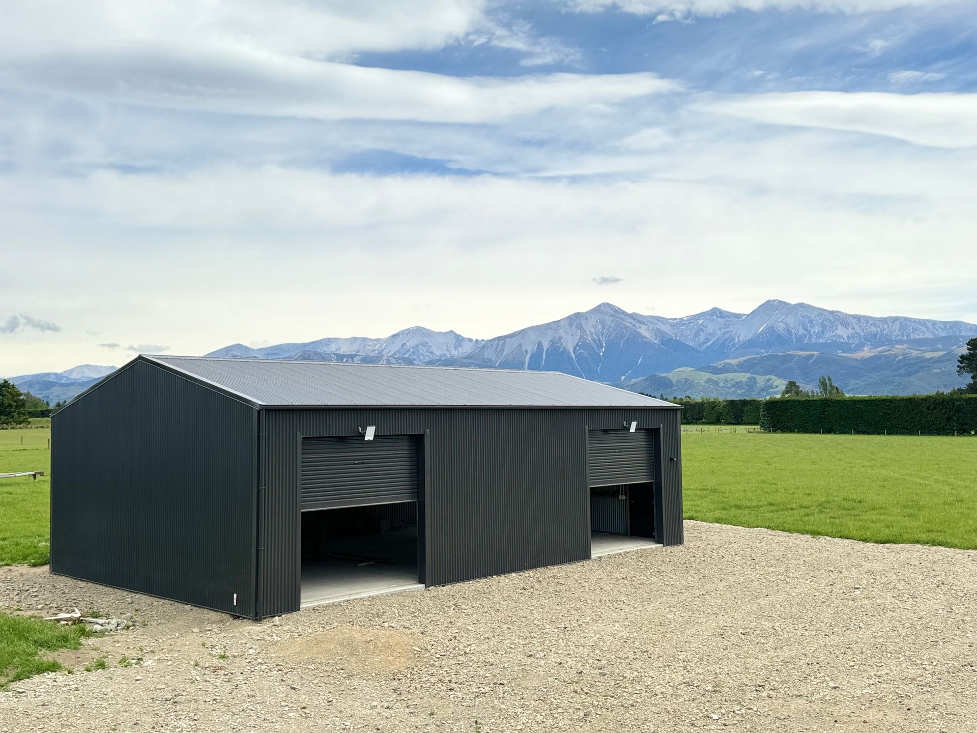 A black garage with two garage doors is sitting in the middle of a grassy field with mountains in the background.