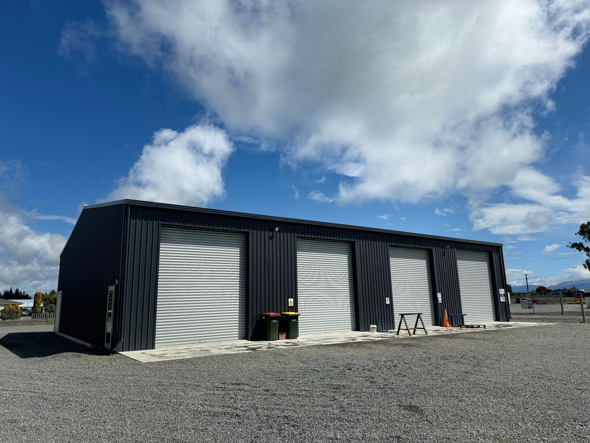 A large metal building with three garage doors is sitting on top of a gravel lot.