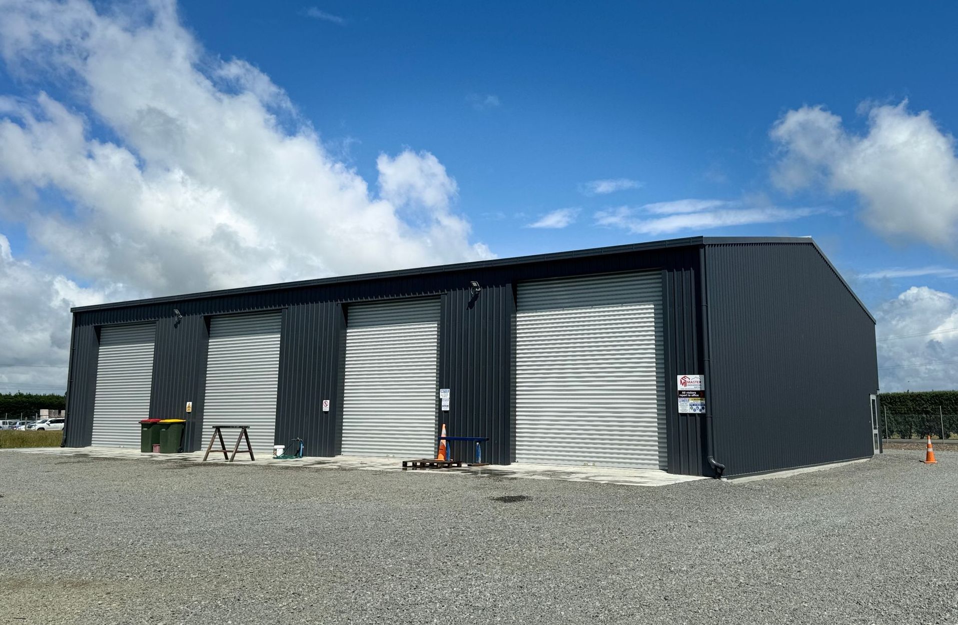 A large metal building with three garage doors is sitting on top of a gravel lot.