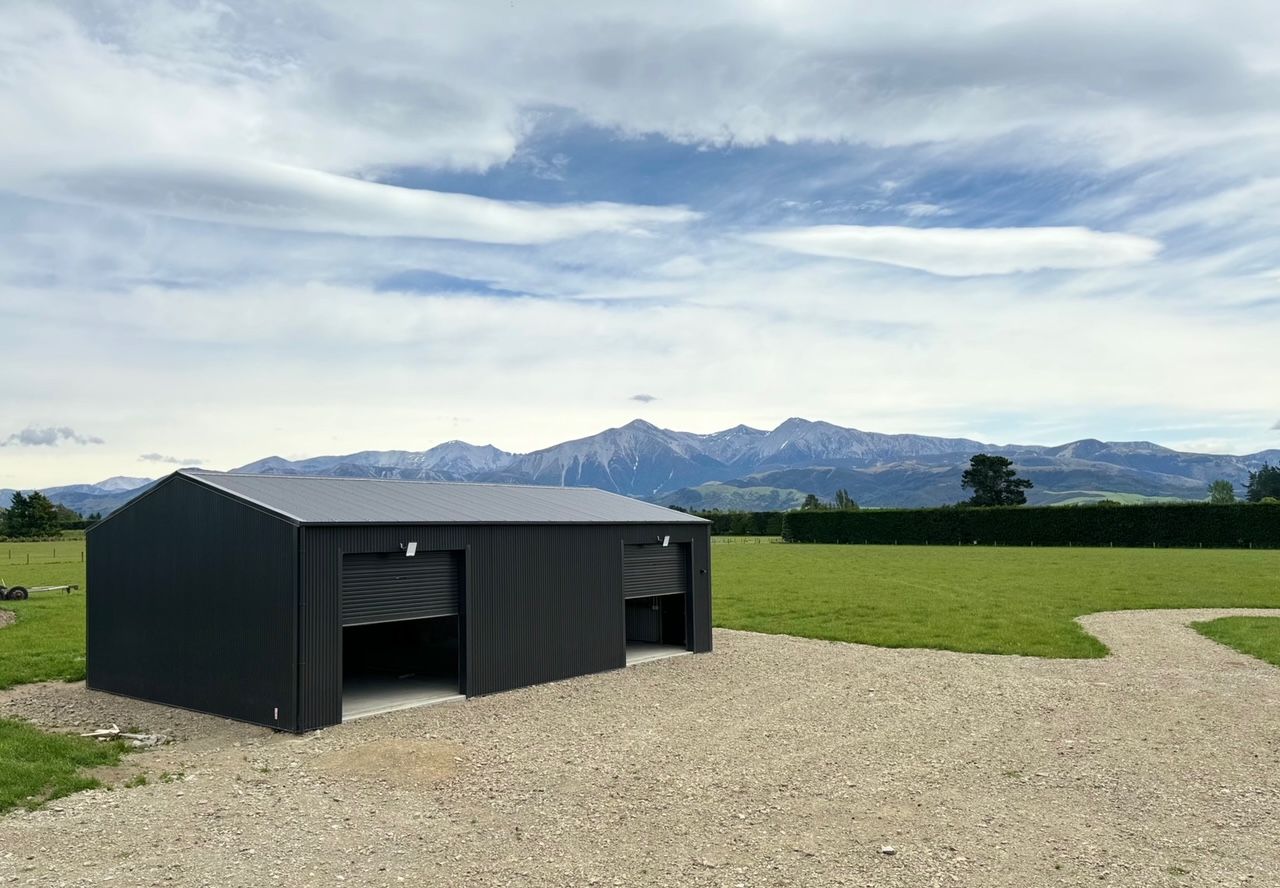 A black shed is sitting in the middle of a grassy field with mountains in the background.
