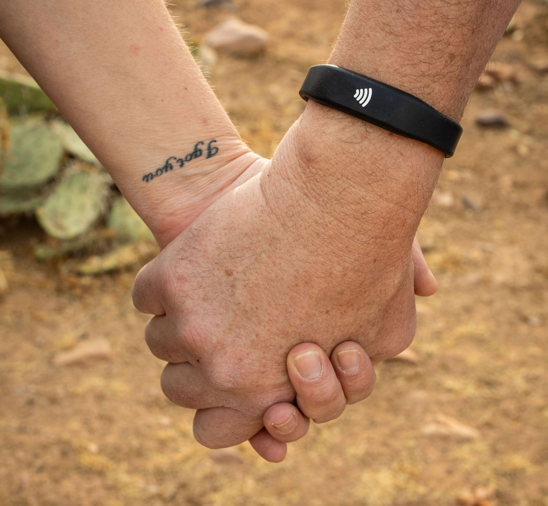 Two people holding hands, one with a black wristband, against a dirt background
