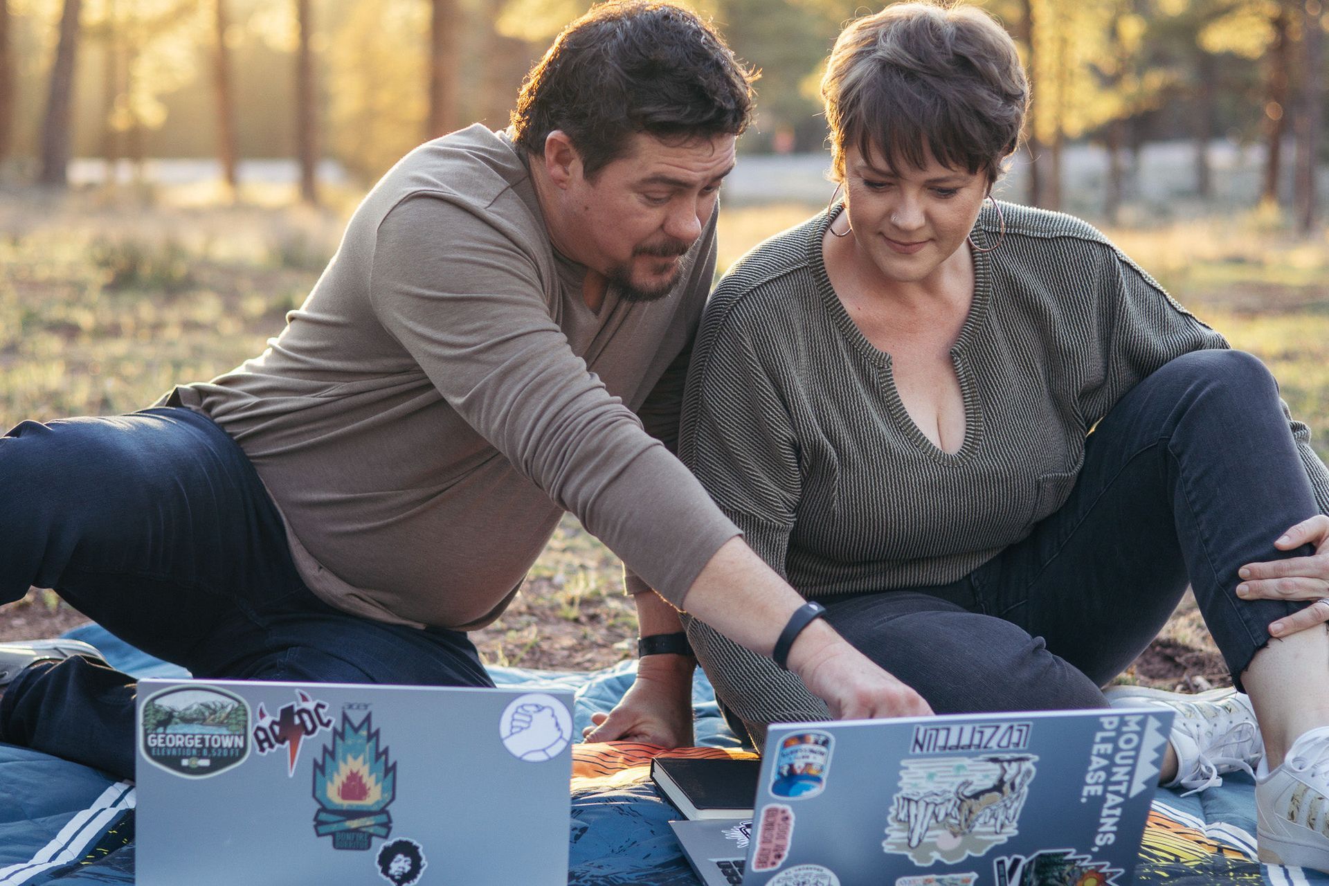Two people sitting outdoors on blankets, working on laptops in a park.